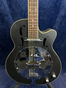 Black electric guitar with a round, shiny metal resonator plate and two control knobs, shown against a dark blue fabric background.
