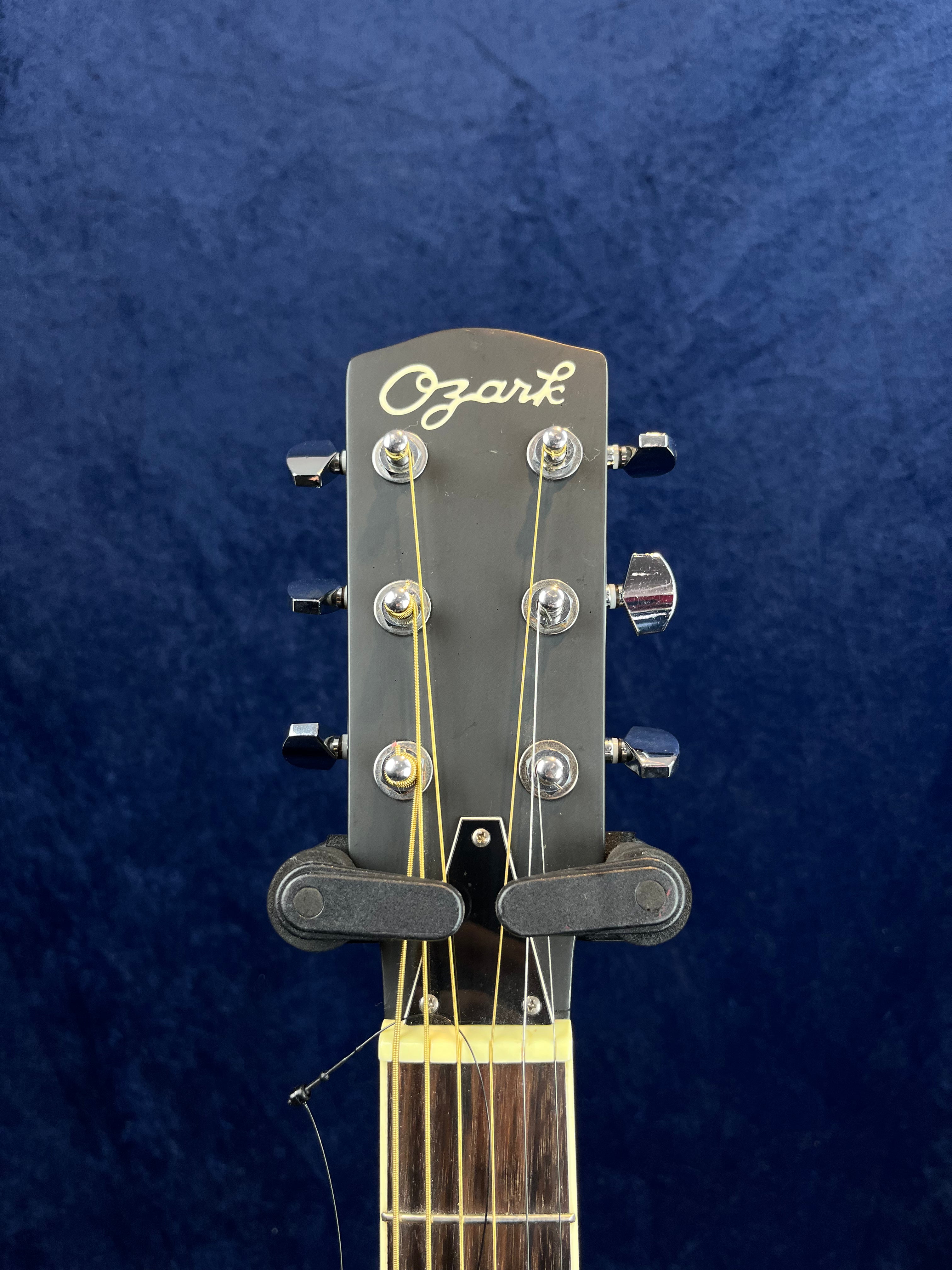 Tête de guitare électrique Ozark noire avec six mécaniques argentées, deux supports en plastique noir tenant le manche, fond bleu uni.