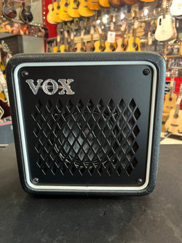 Petit ampli noir VOX avec grille en losanges sur la façade, posé sur une table, guitares accrochées en arrière-plan.