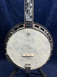 Five-string banjo with a white drumhead, silver hardware, and ornate inlay designs on the fingerboard, displayed against a dark blue background.