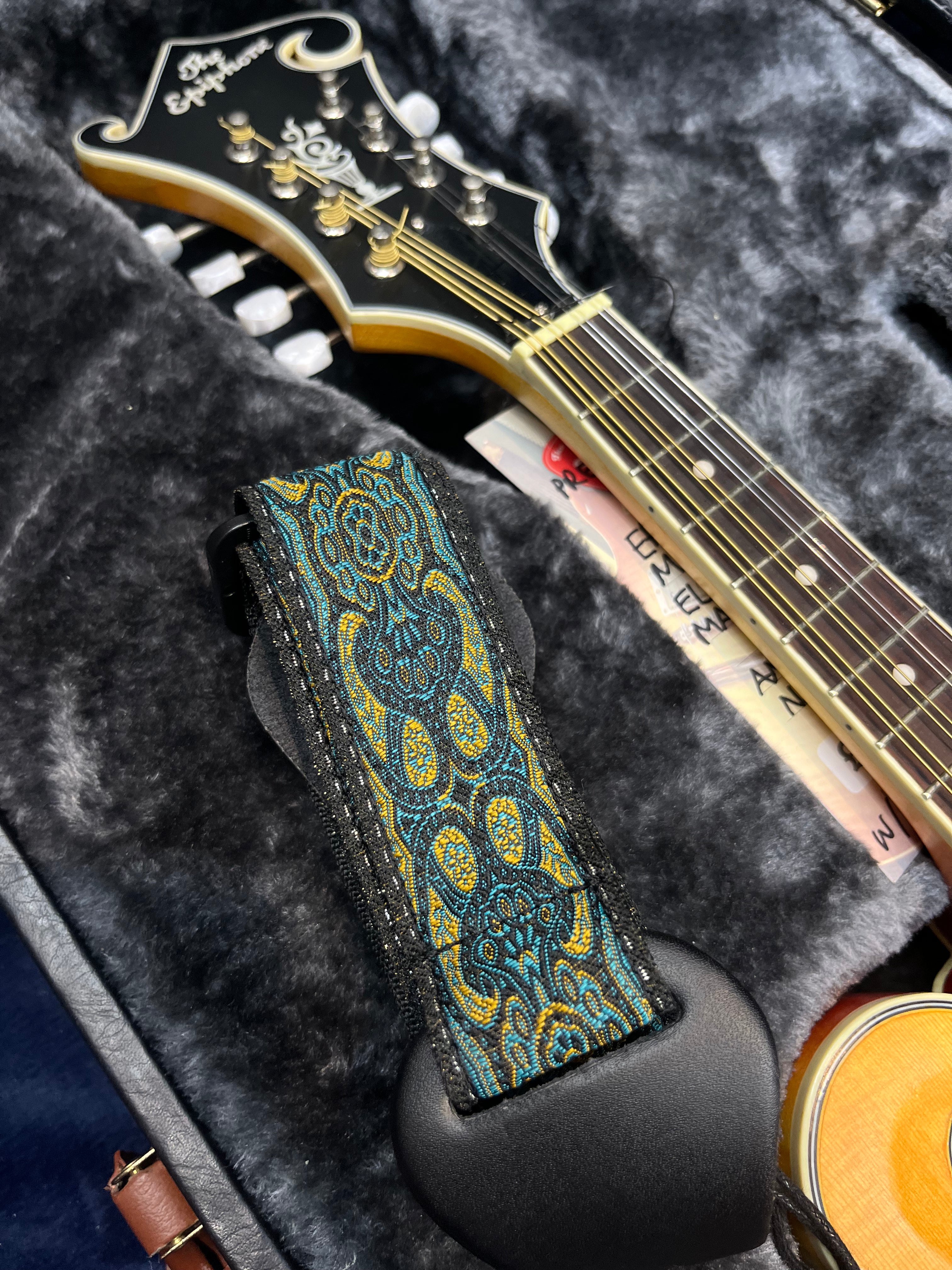 Tête de mandoline noire avec motif doré, étui molletonné gris, sangle colorée bleu et jaune à motifs, touches et cordes visibles.