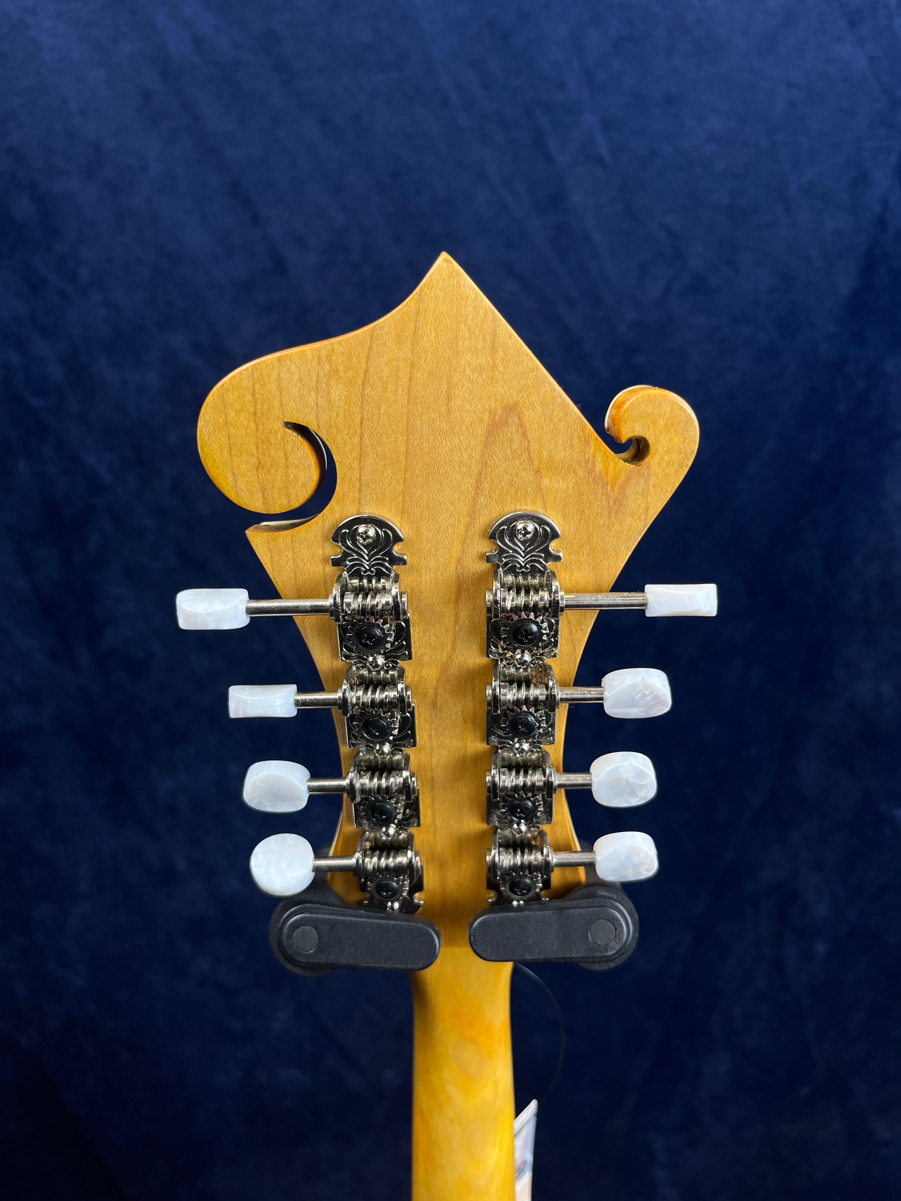 Tête de mandoline vue de dos, bois clair sculpté, huit mécaniques argentées avec boutons blancs, fond bleu.