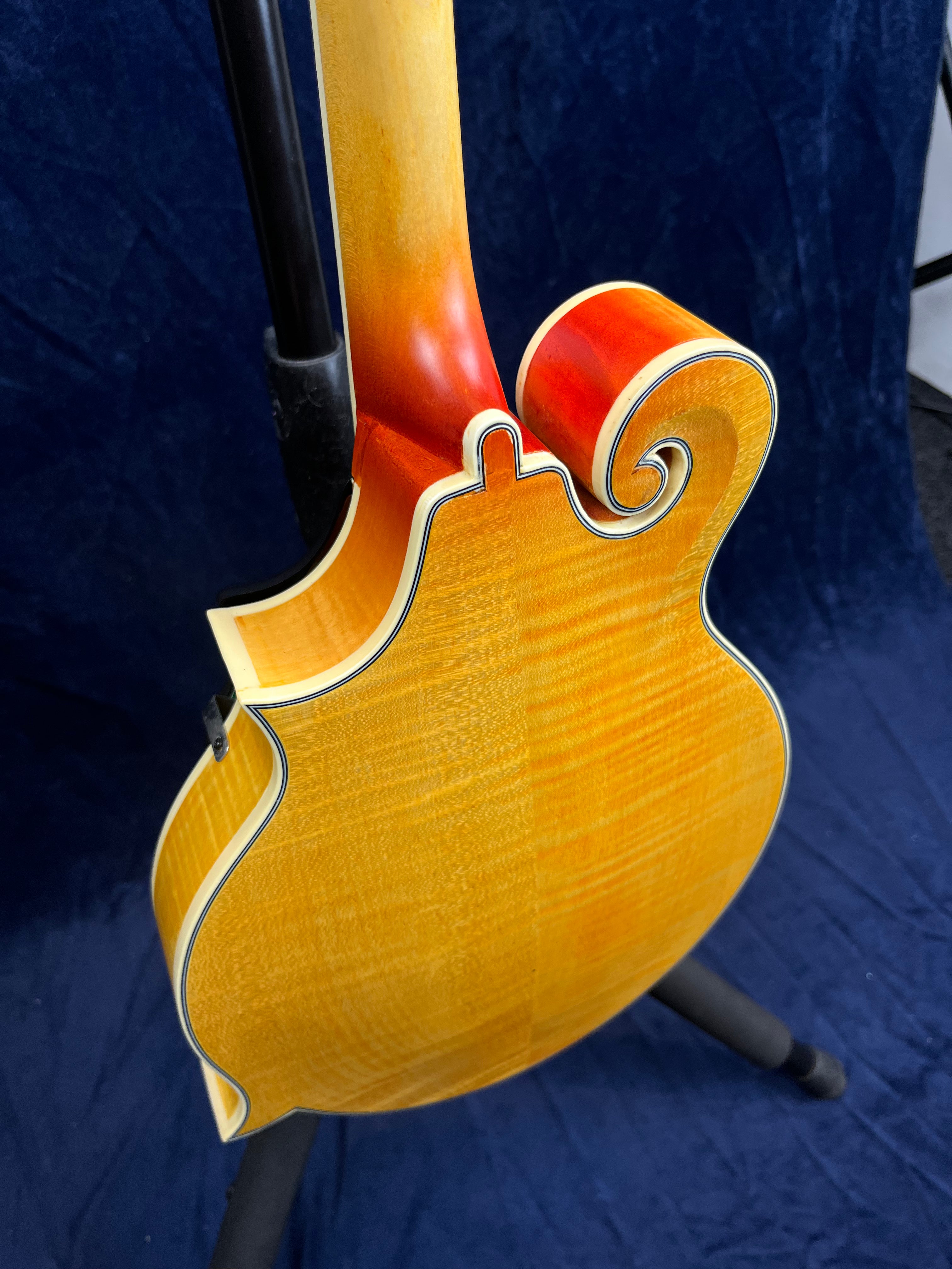 Mandoline à dos ondé jaune avec bordures blanches et noires, forme sculptée, manche jaune clair, fond dégradé orangé, posée sur un support.