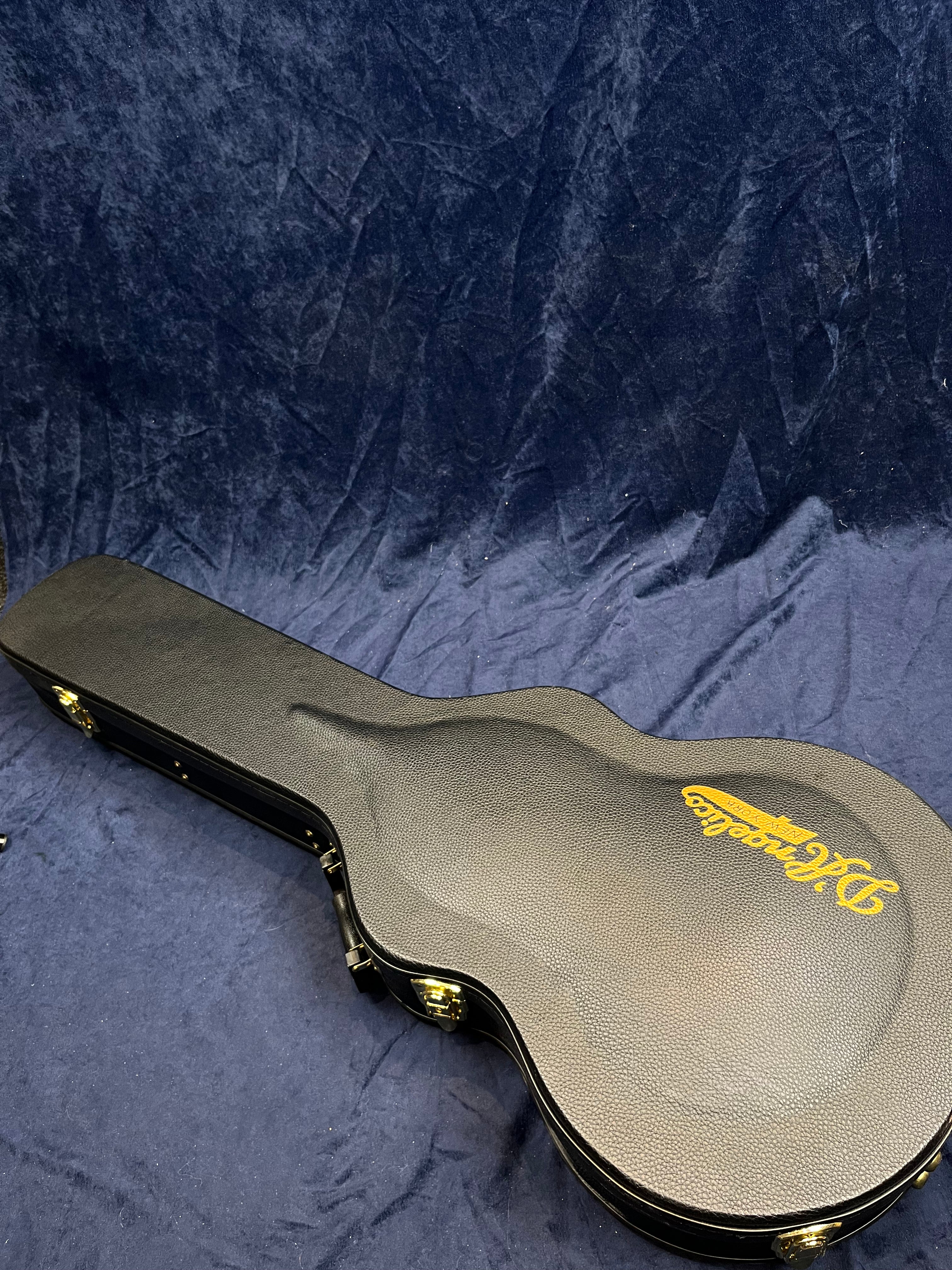 Hard guitar case with textured black surface, gold D’Angelico logo, and metal latches resting on a dark, crumpled fabric background.