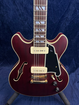 Semi-hollow electric guitar with deep red finish, cream pickups, gold hardware, black pickguard, dual f-holes, and block inlays on the fretboard.