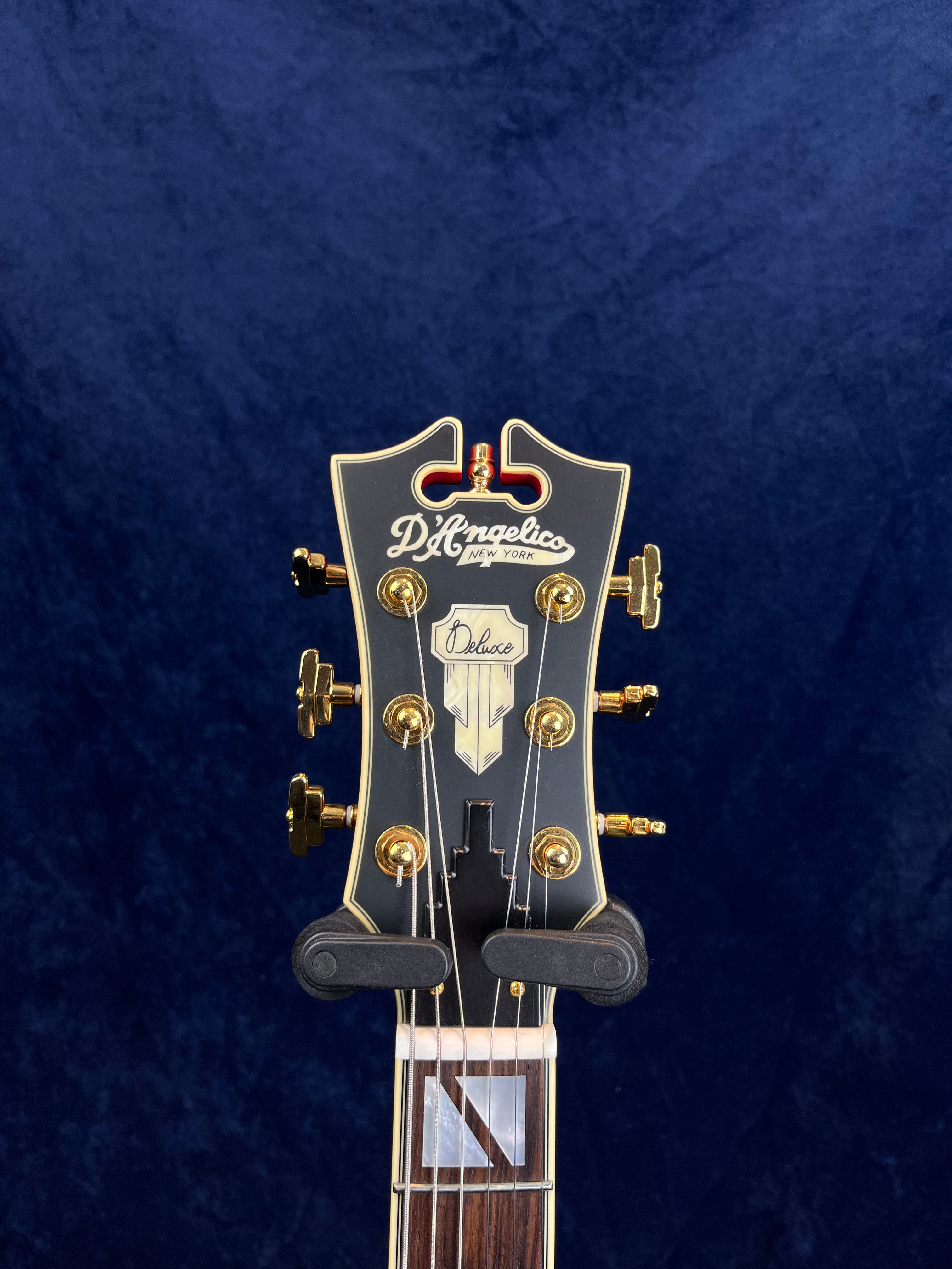 D’Angelico electric guitar headstock with gold tuners, black finish, inlaid logo, and “Deluxe” label above decorative truss cover.