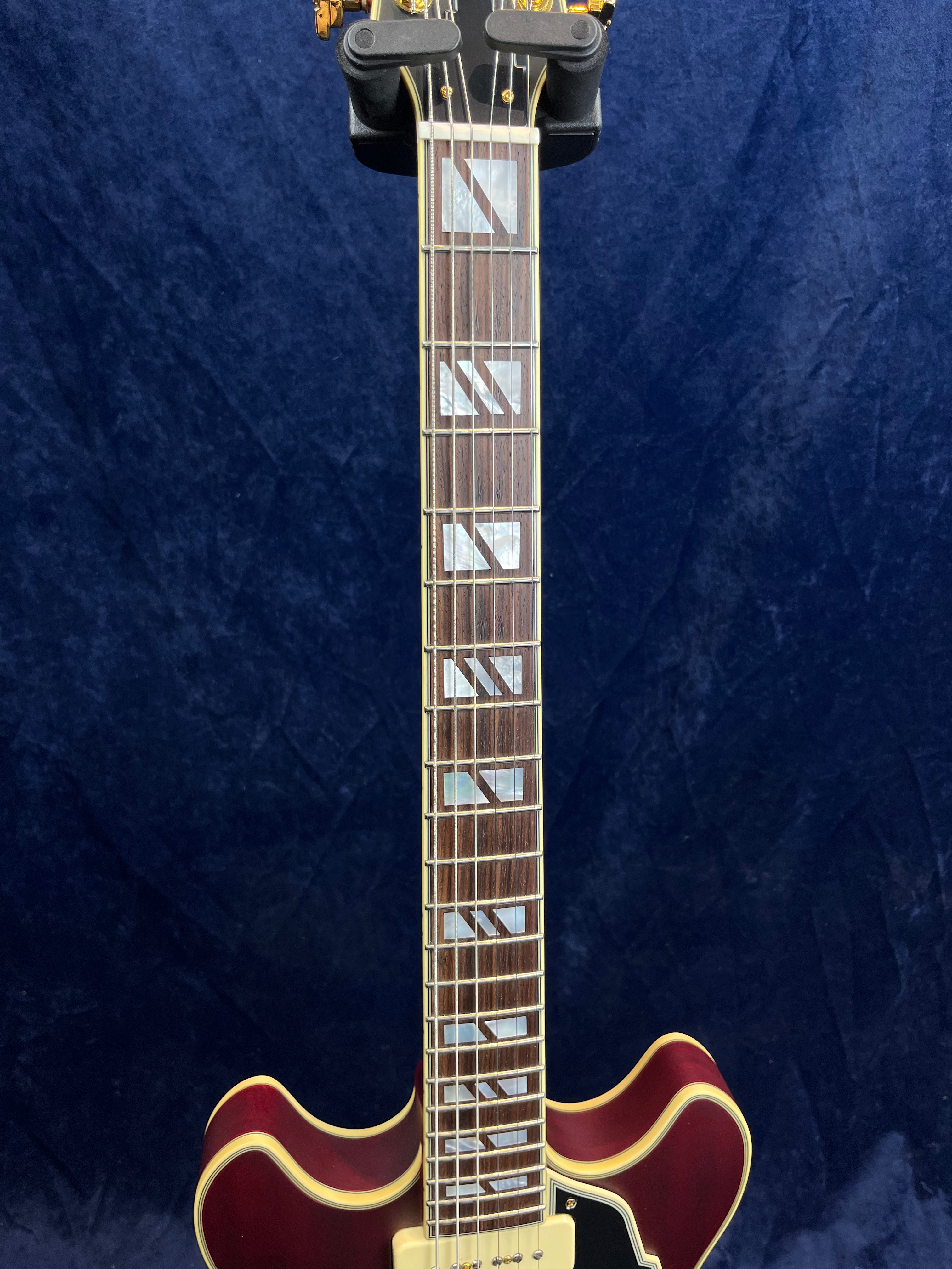 Cherry red electric guitar with cream binding, geometric fretboard inlays, and double cutaway body against a dark blue background.