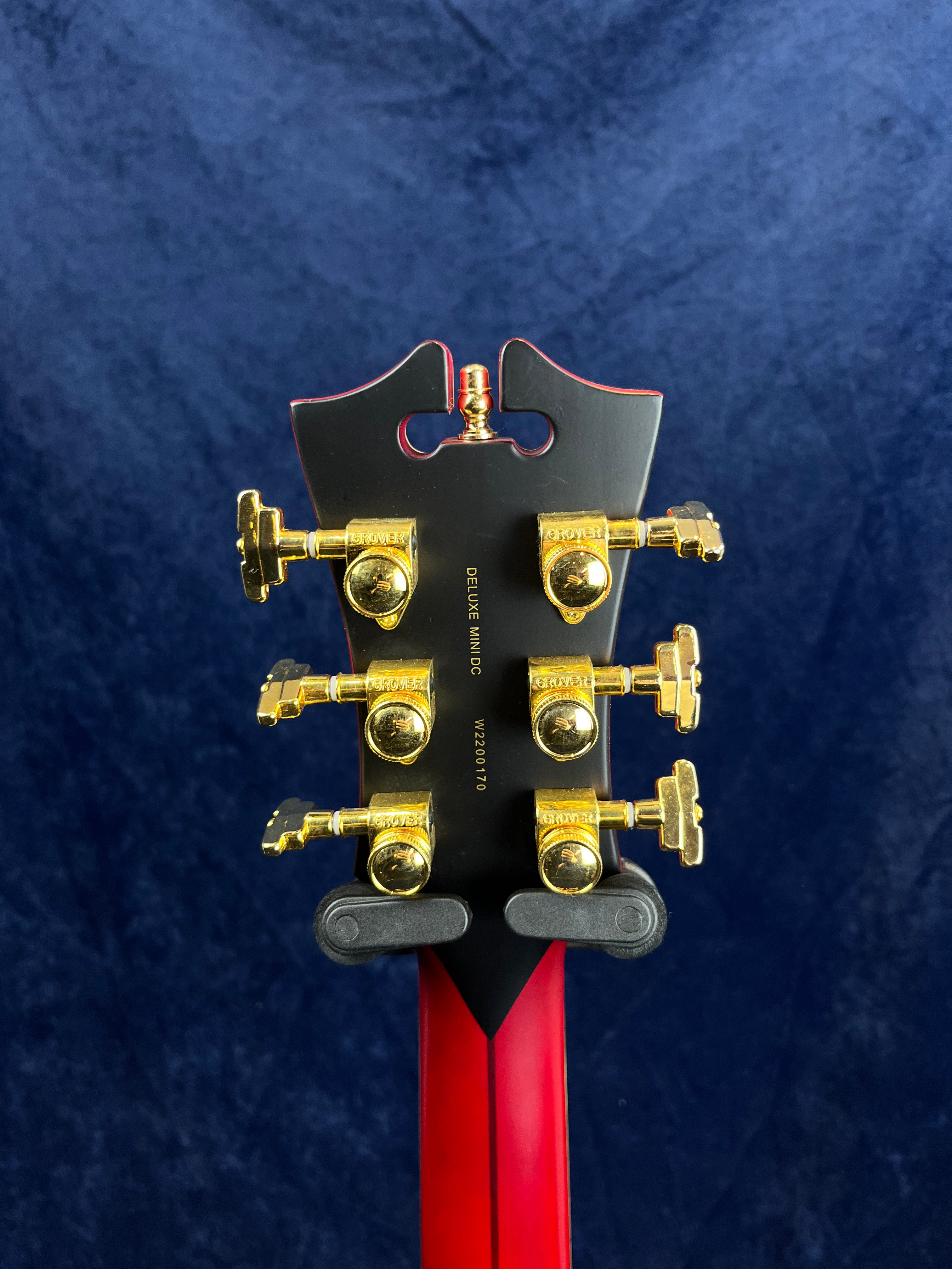 Electric guitar headstock in black and red with six gold tuning pegs, “Grover” branding, and “DELUXE” and serial number text visible.