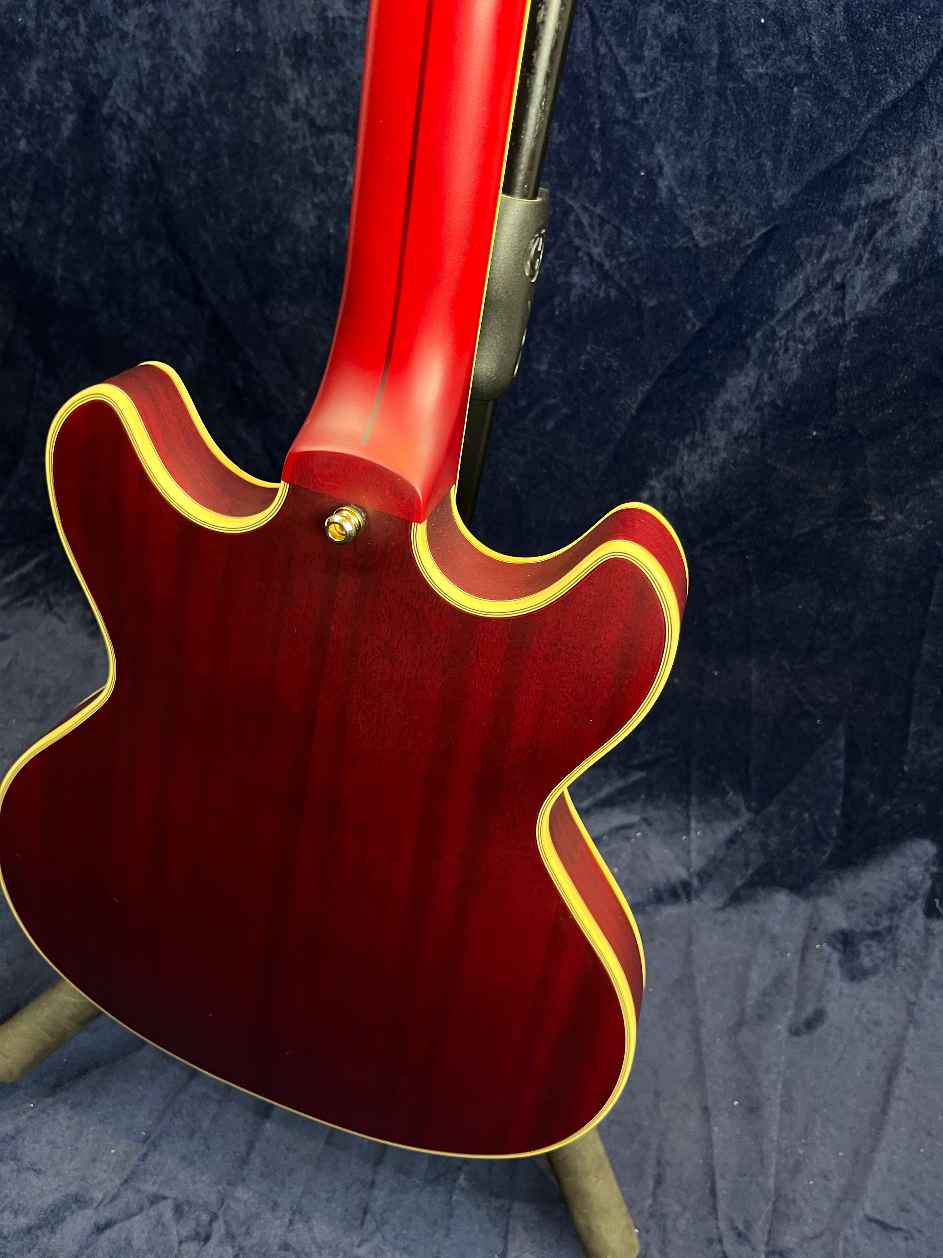 Deep red semi-hollow electric guitar viewed from the back, with cream binding outlining the body and neck, resting on a stand against a dark backdrop.