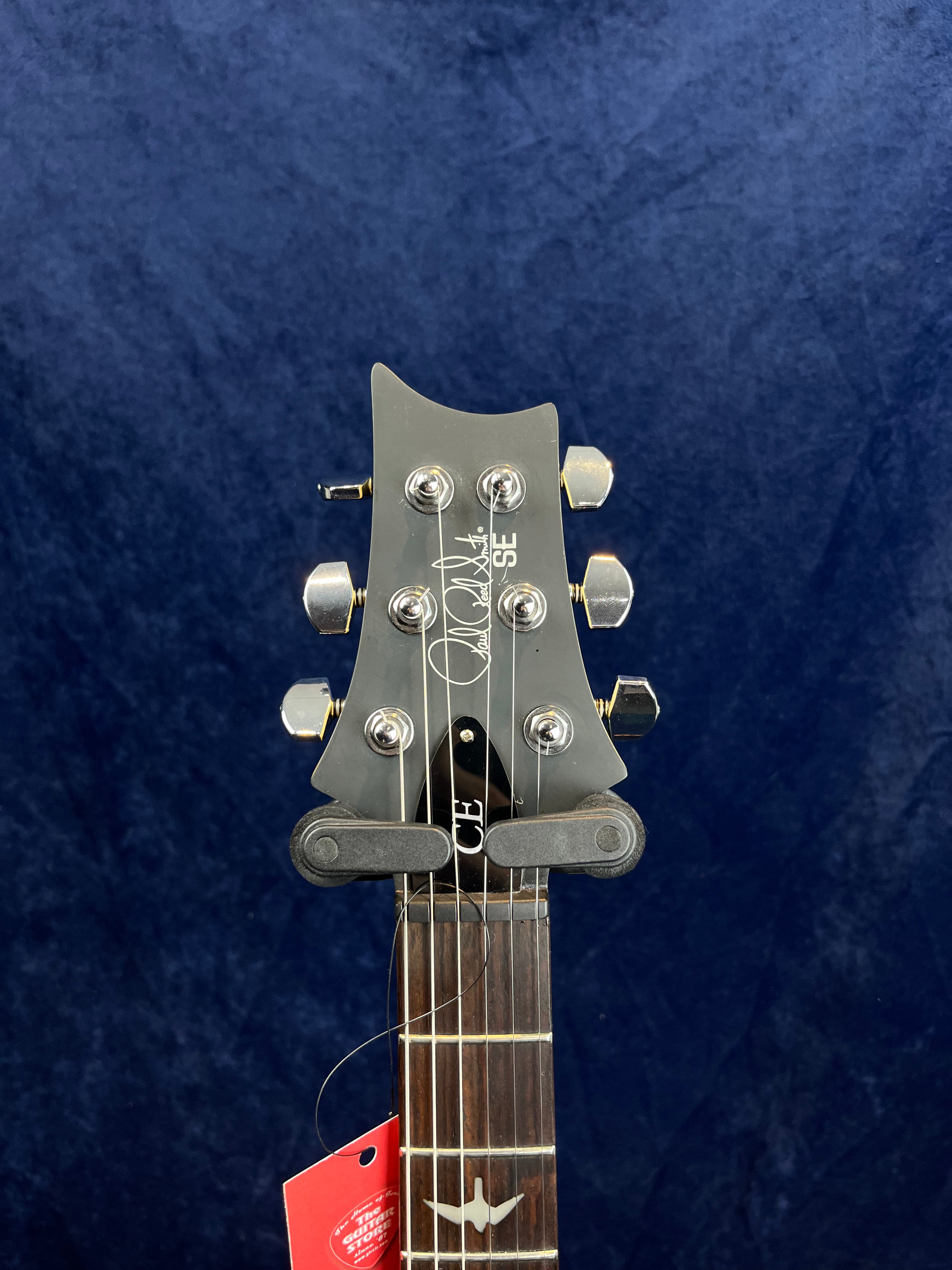 Tête de guitare électrique PRS SE noire avec six mécaniques chromées et logo signature blanc, manche avec repère en forme d’oiseau.