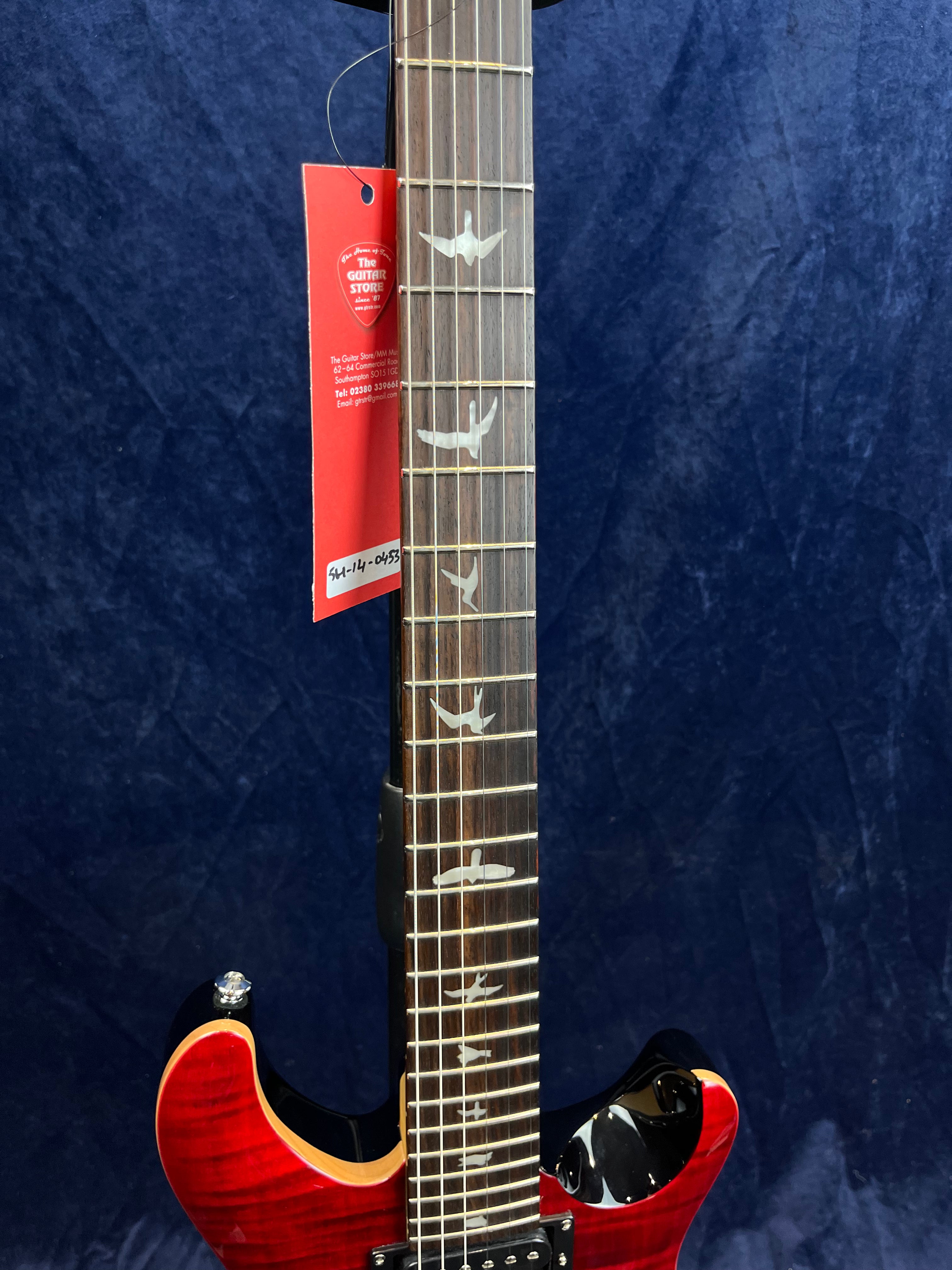 Guitare électrique avec manche orné d'incrustations en forme d’oiseaux, corps rouge brillant et étiquette rouge accrochée à une corde.