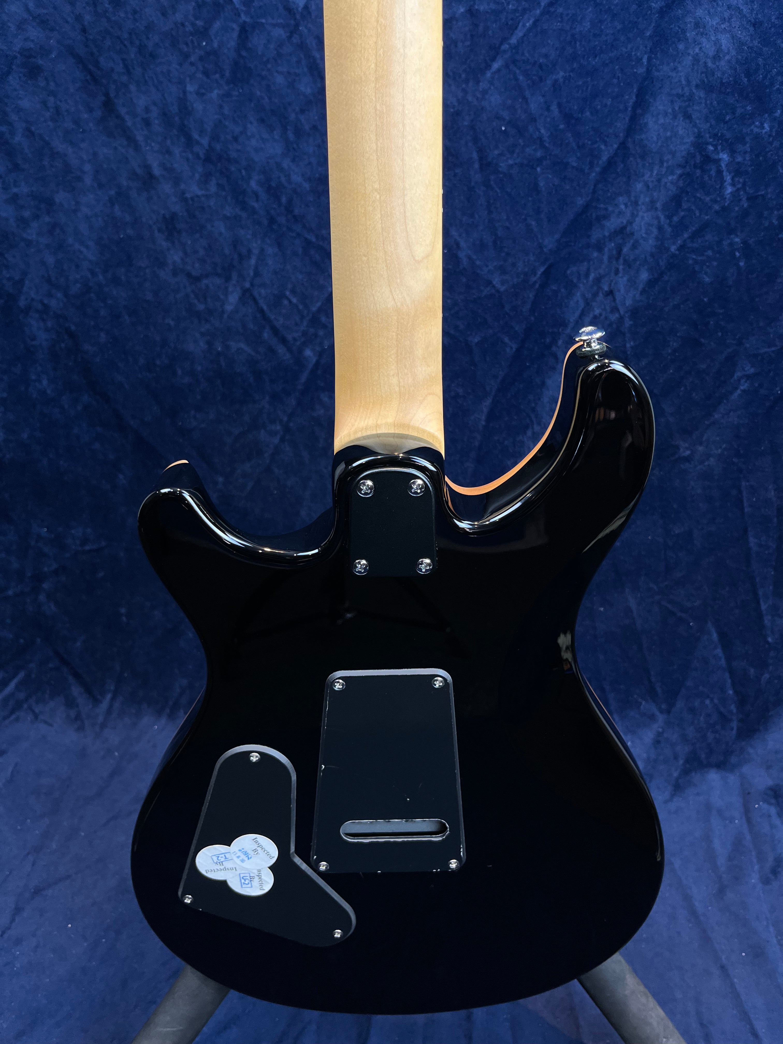 Guitare électrique noire vue de dos, manche érable clair, plaques d’accès vissées et autocollants sur la plaque inférieure.