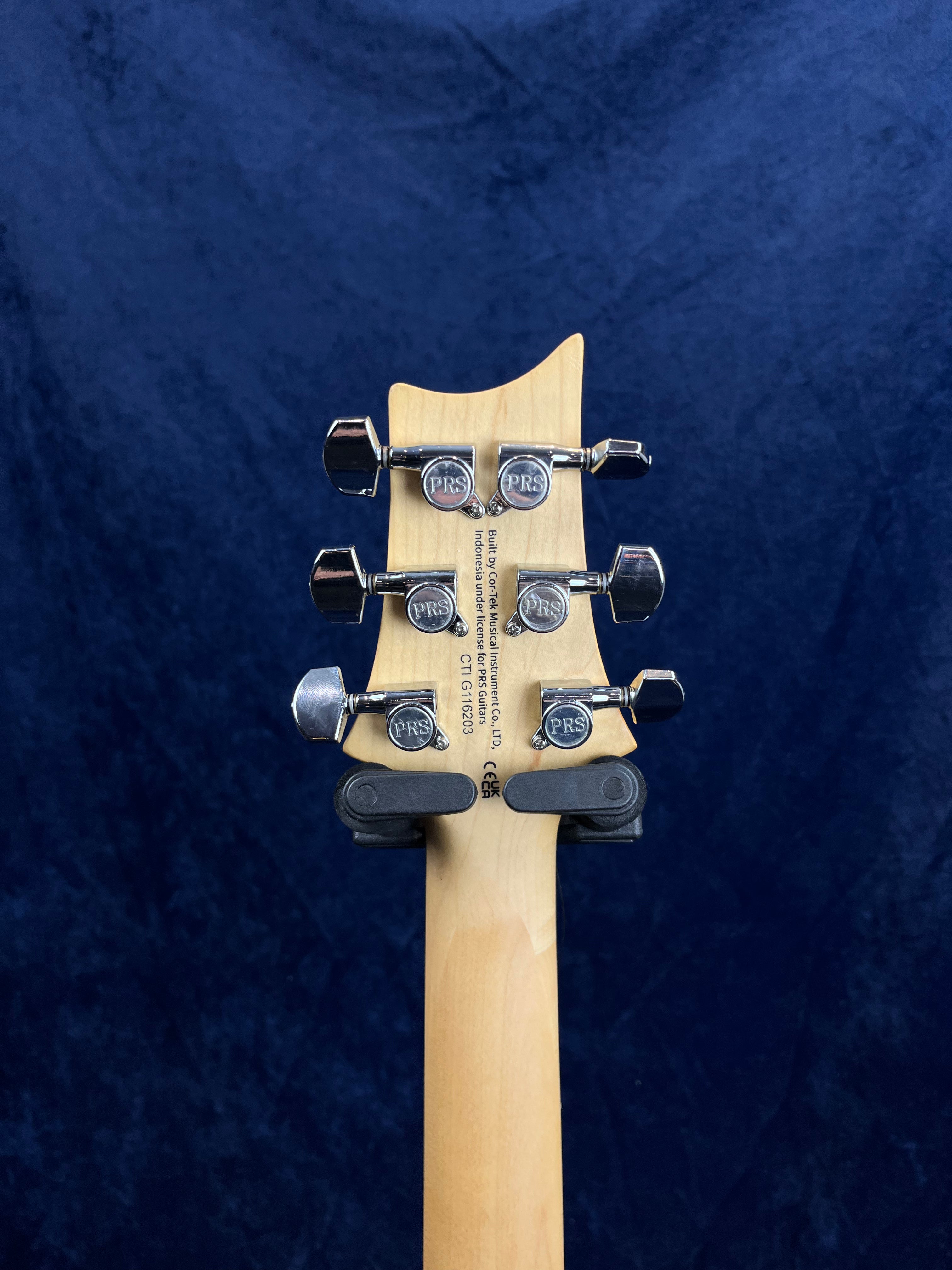 Tête d’une guitare électrique en bois clair avec six mécaniques argentées marquées « PRS », fond bleu sombre uniforme.