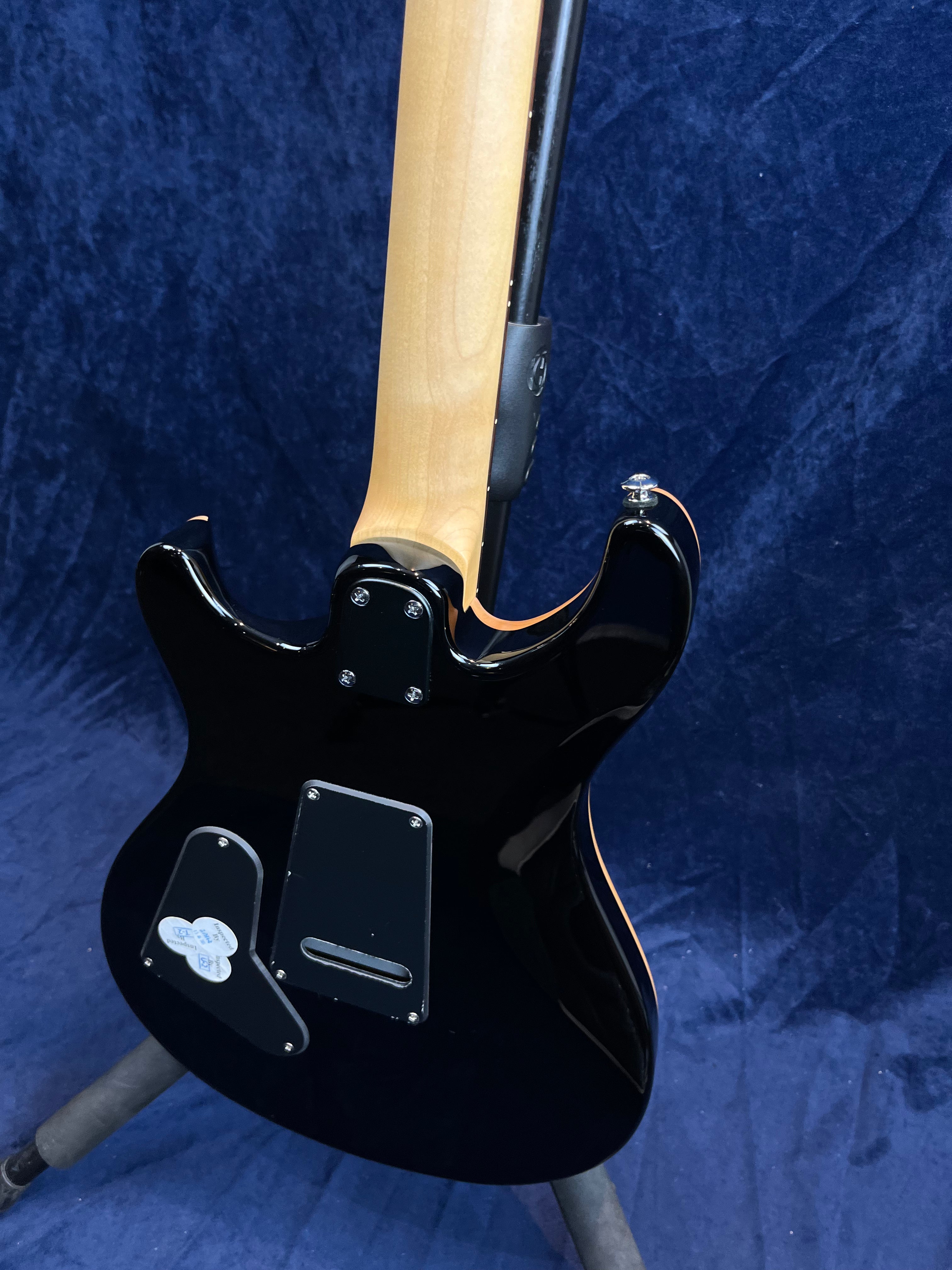 Guitare électrique noire vue de dos, manche en bois clair, plaques d’accès fixées à l’arrière du corps, deux médiators coincés sur une plaque.