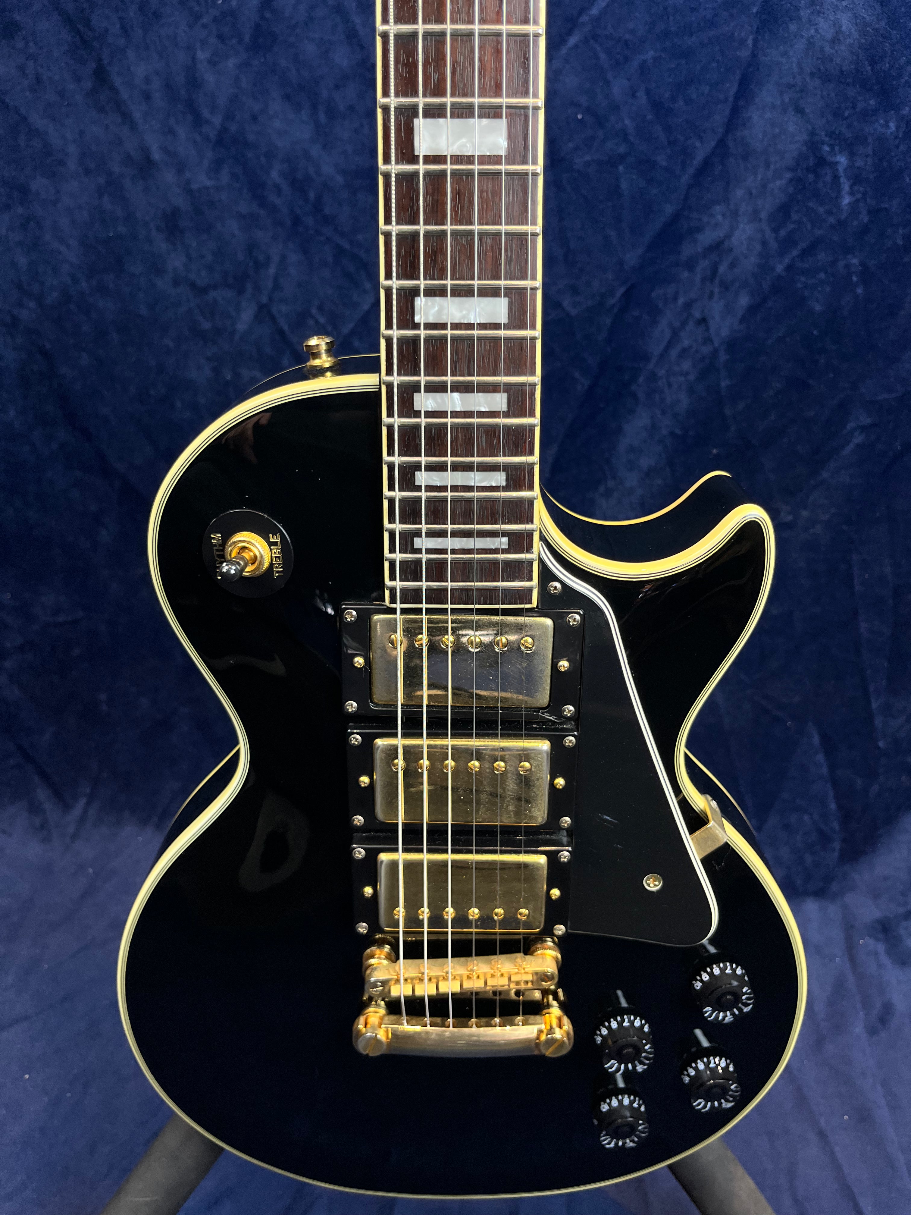 Black electric guitar with three gold humbucker pickups, gold hardware, four black control knobs, and white trim, shown against a dark blue background.
