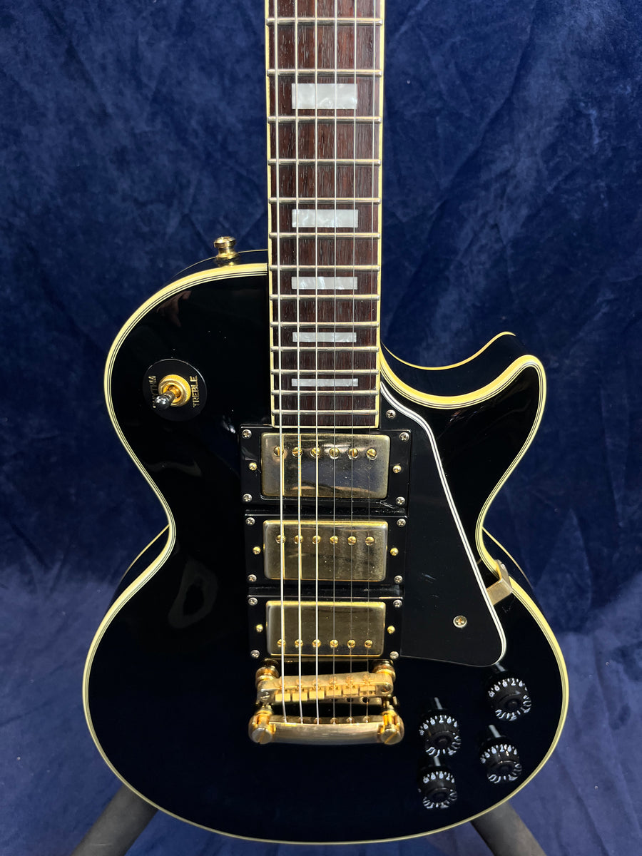 Black electric guitar with three gold humbucker pickups, gold hardware, four black control knobs, and white trim, shown against a dark blue background.