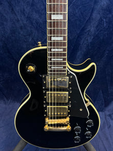 Black electric guitar with three gold humbucker pickups, gold hardware, four black control knobs, and white trim, shown against a dark blue background.
