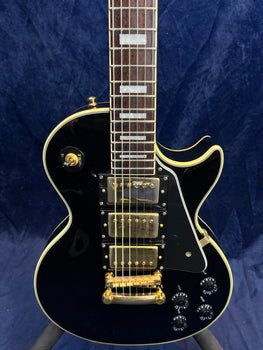 Black electric guitar with three gold humbucker pickups, gold hardware, four black control knobs, and white trim, shown against a dark blue background.