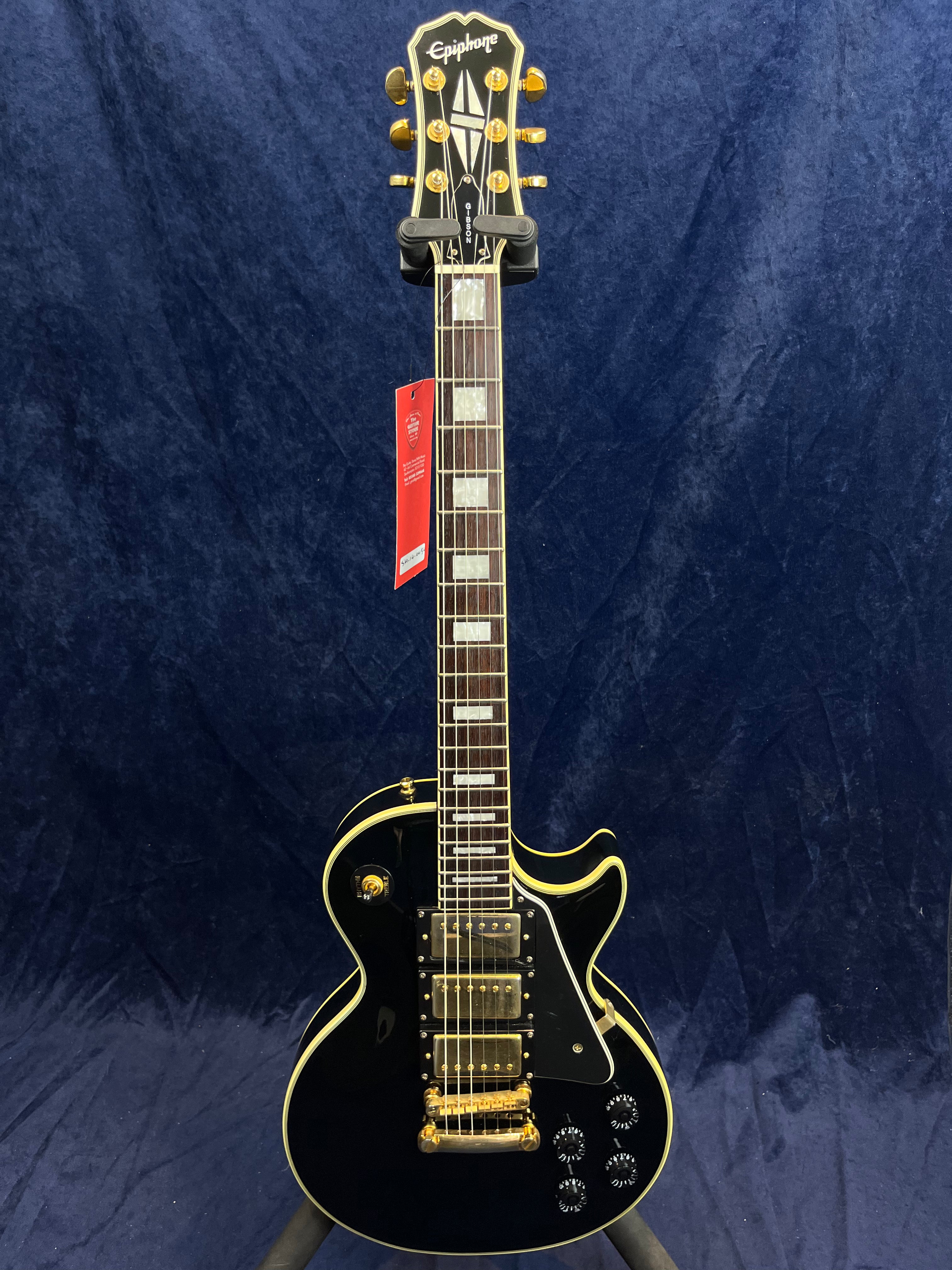 Black Epiphone electric guitar with gold hardware, three pickups, cream binding, rectangular inlays, and a red tag hanging from the neck.