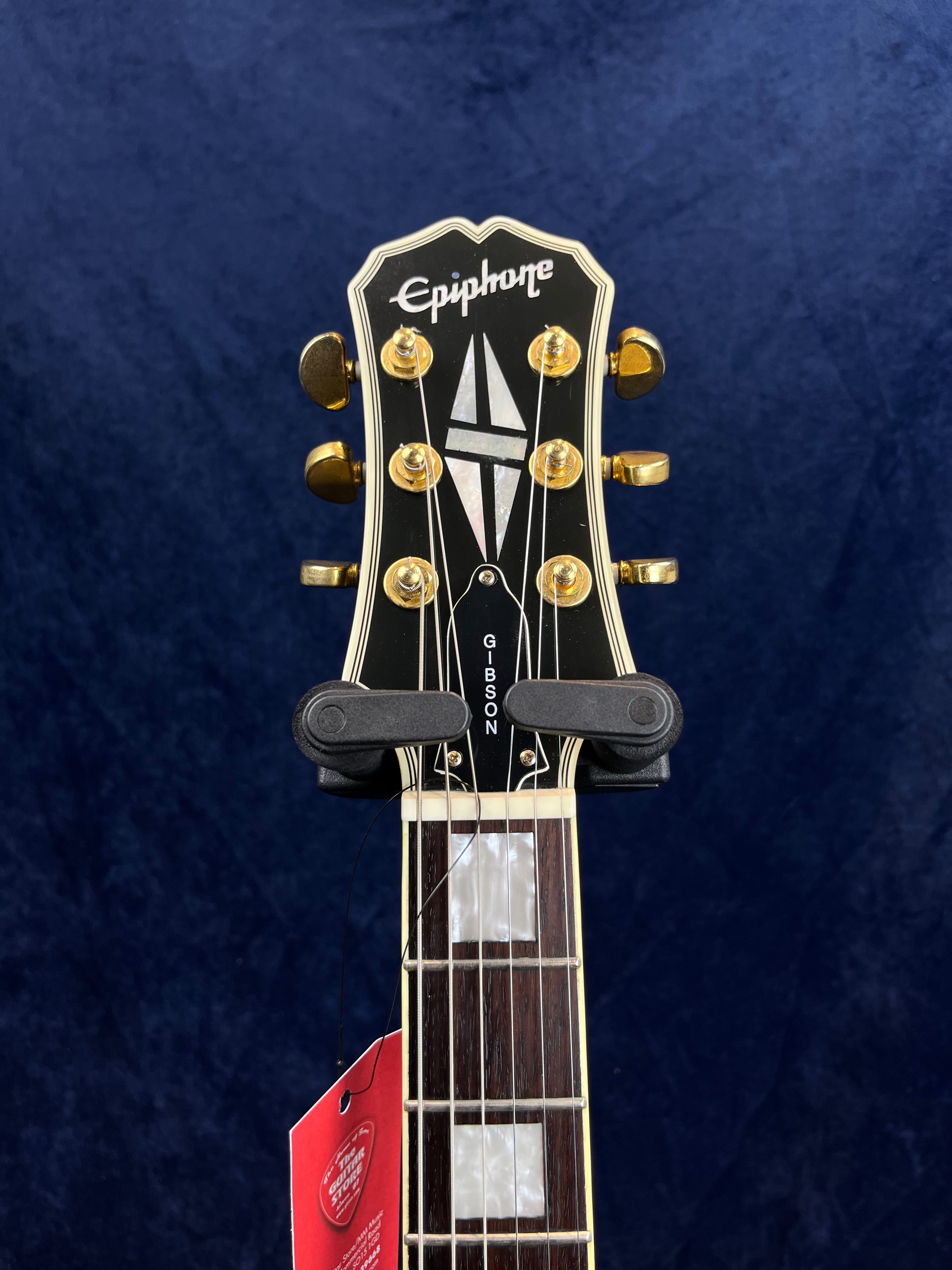 Epiphone electric guitar headstock with gold tuners, mother of pearl inlays, and a truss rod cover labeled Gibson against a dark blue background.