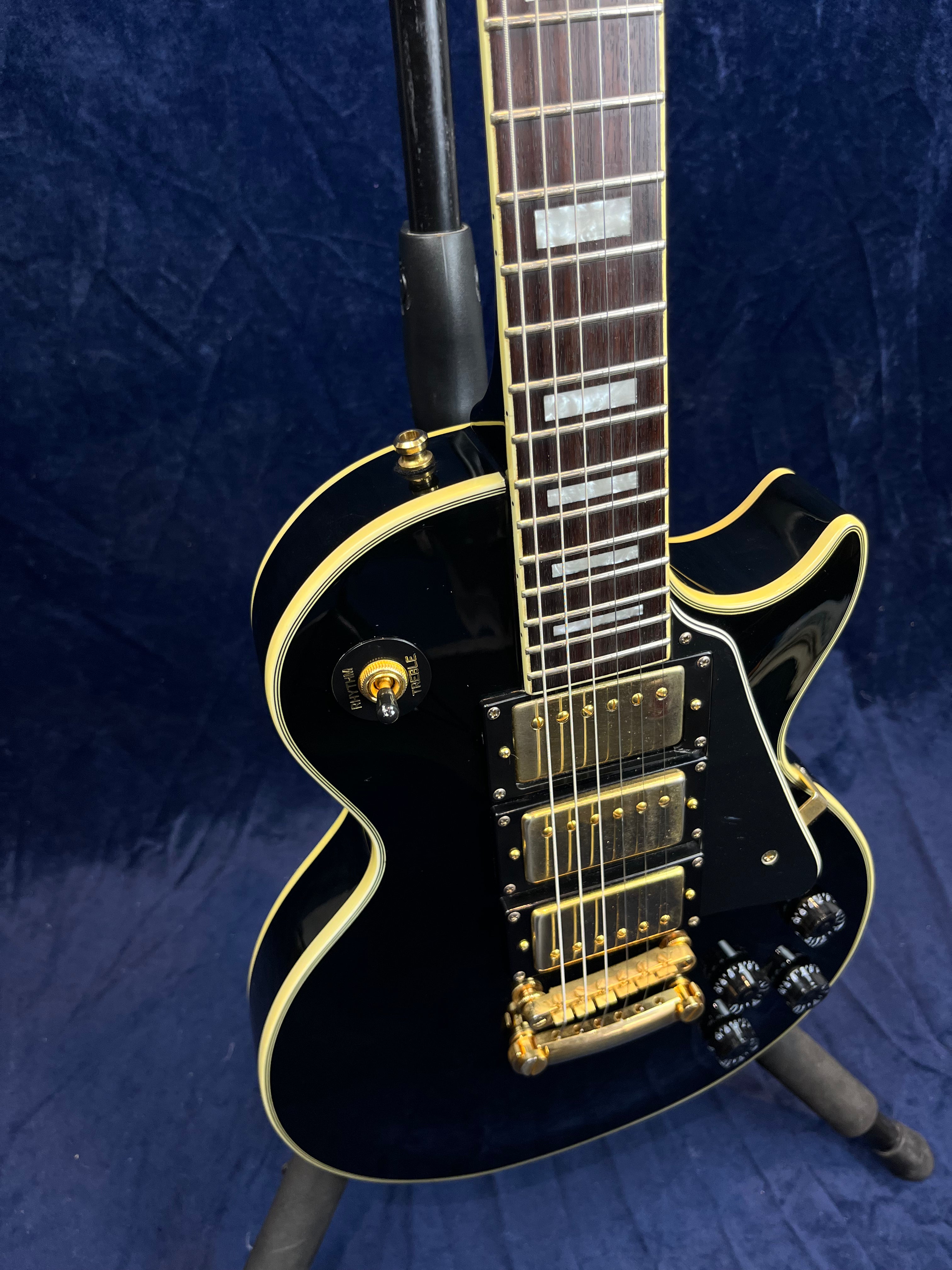 Black electric guitar with gold hardware, three pickups, cream binding, rectangular fretboard inlays, and multiple black control knobs, resting on a stand.