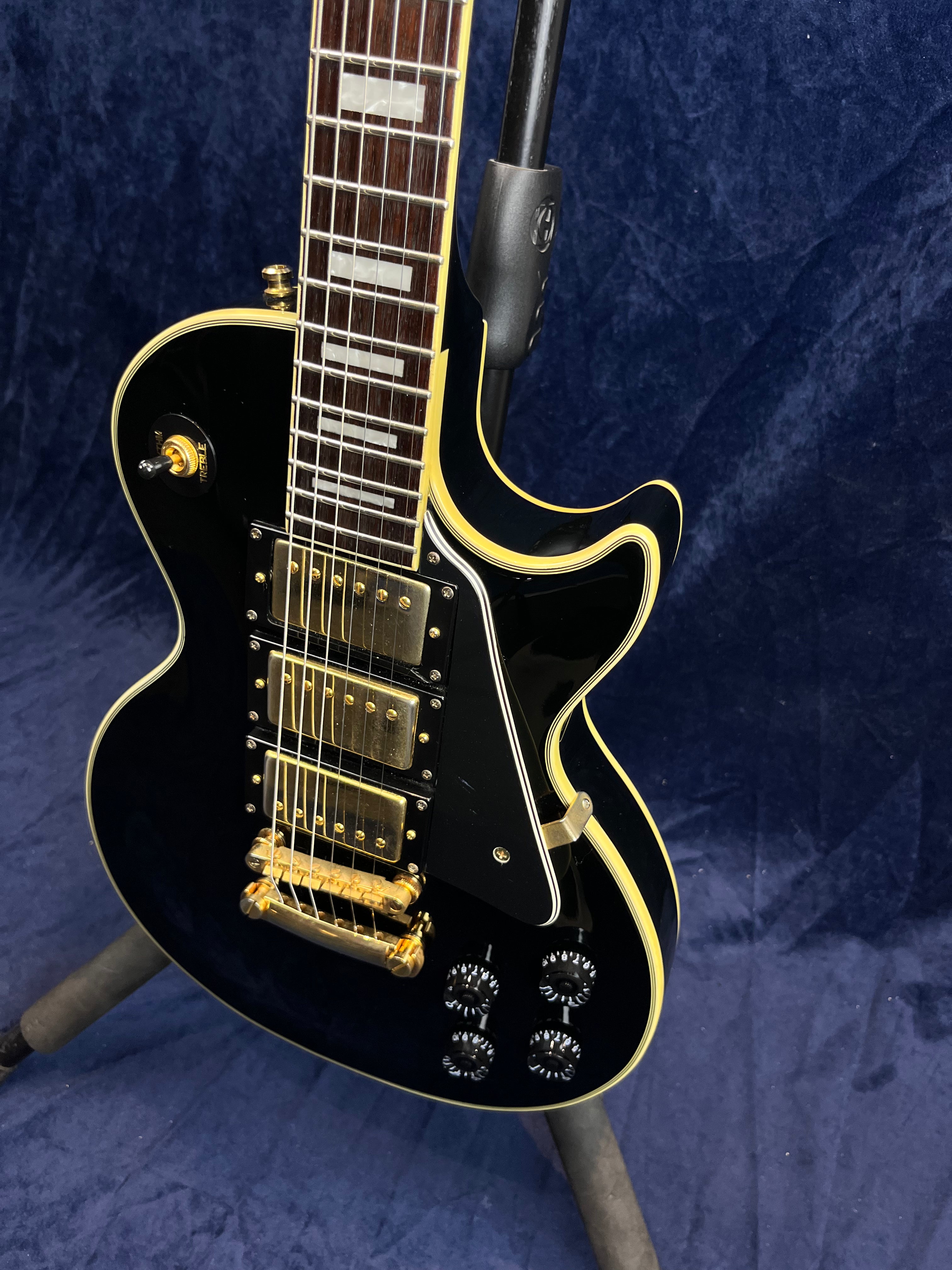 Glossy black electric guitar with gold hardware, three pickups, cream binding, black knobs, and rectangular inlays on the fretboard.