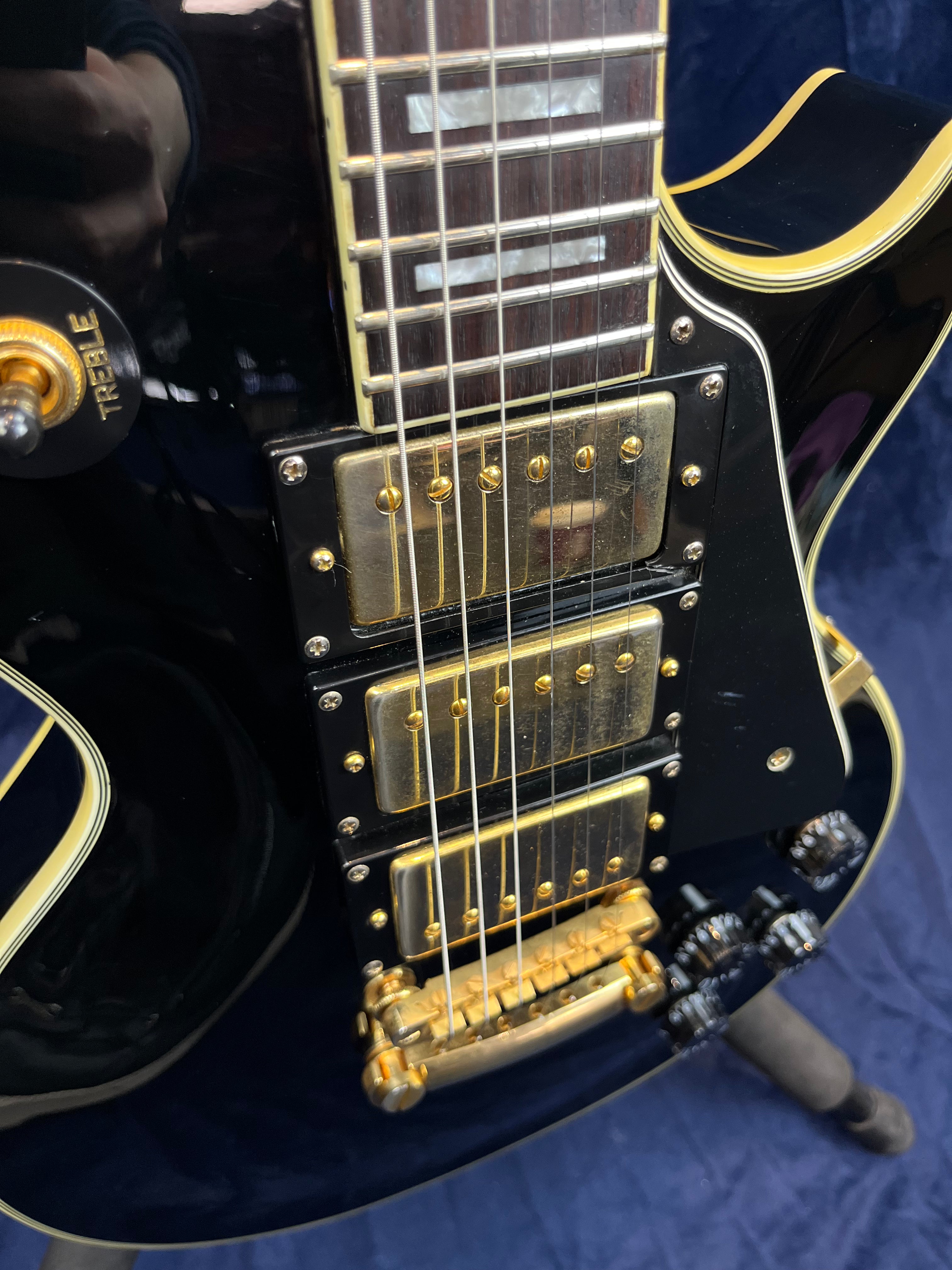 Close-up of a black electric guitar with three gold humbucker pickups, gold hardware, cream binding, and labeled toggle switch reading "Treble."