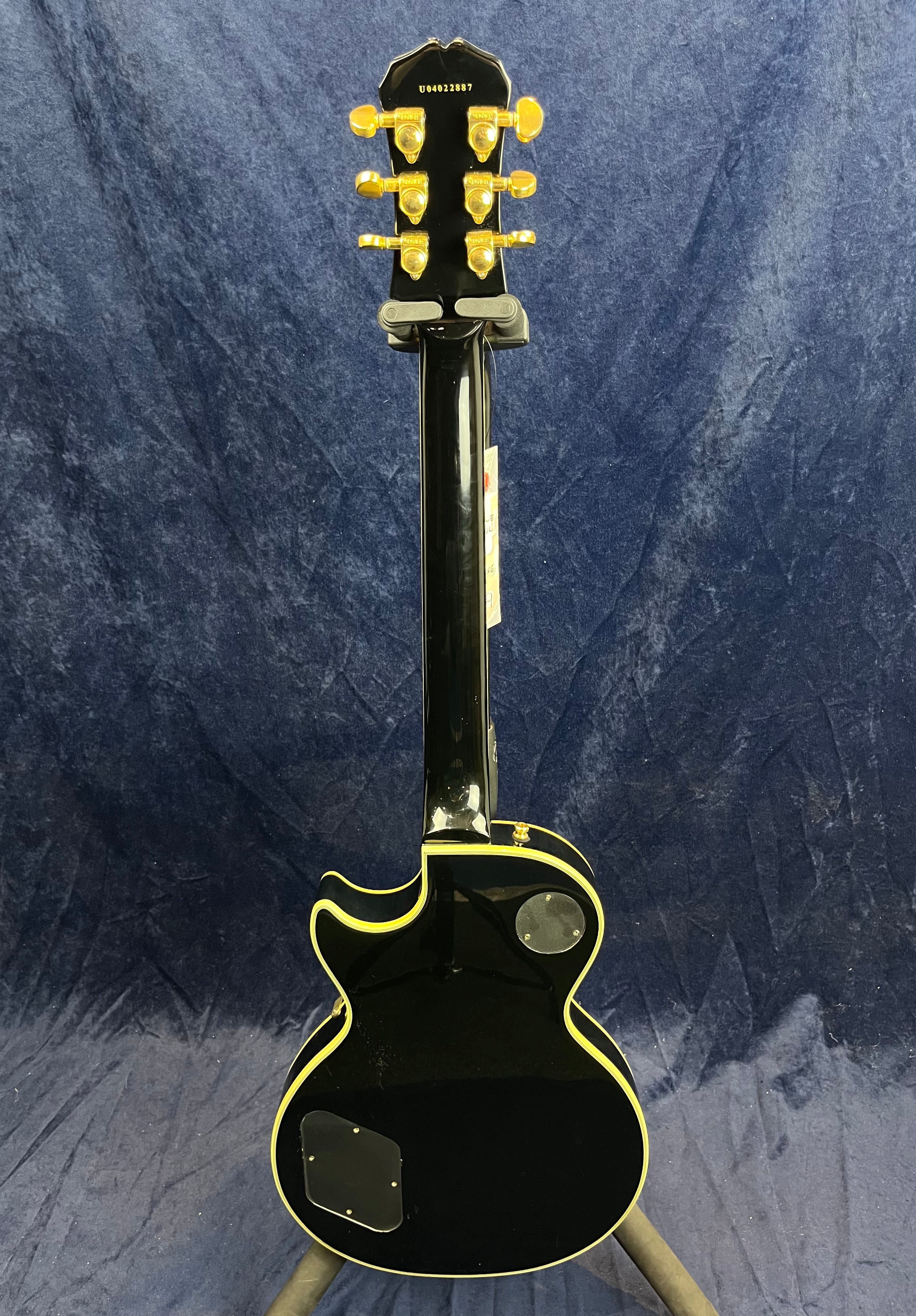 Black electric guitar shown from the back, gold tuning pegs, yellow edge binding, and rear control cavity covers visible against a dark blue background.
