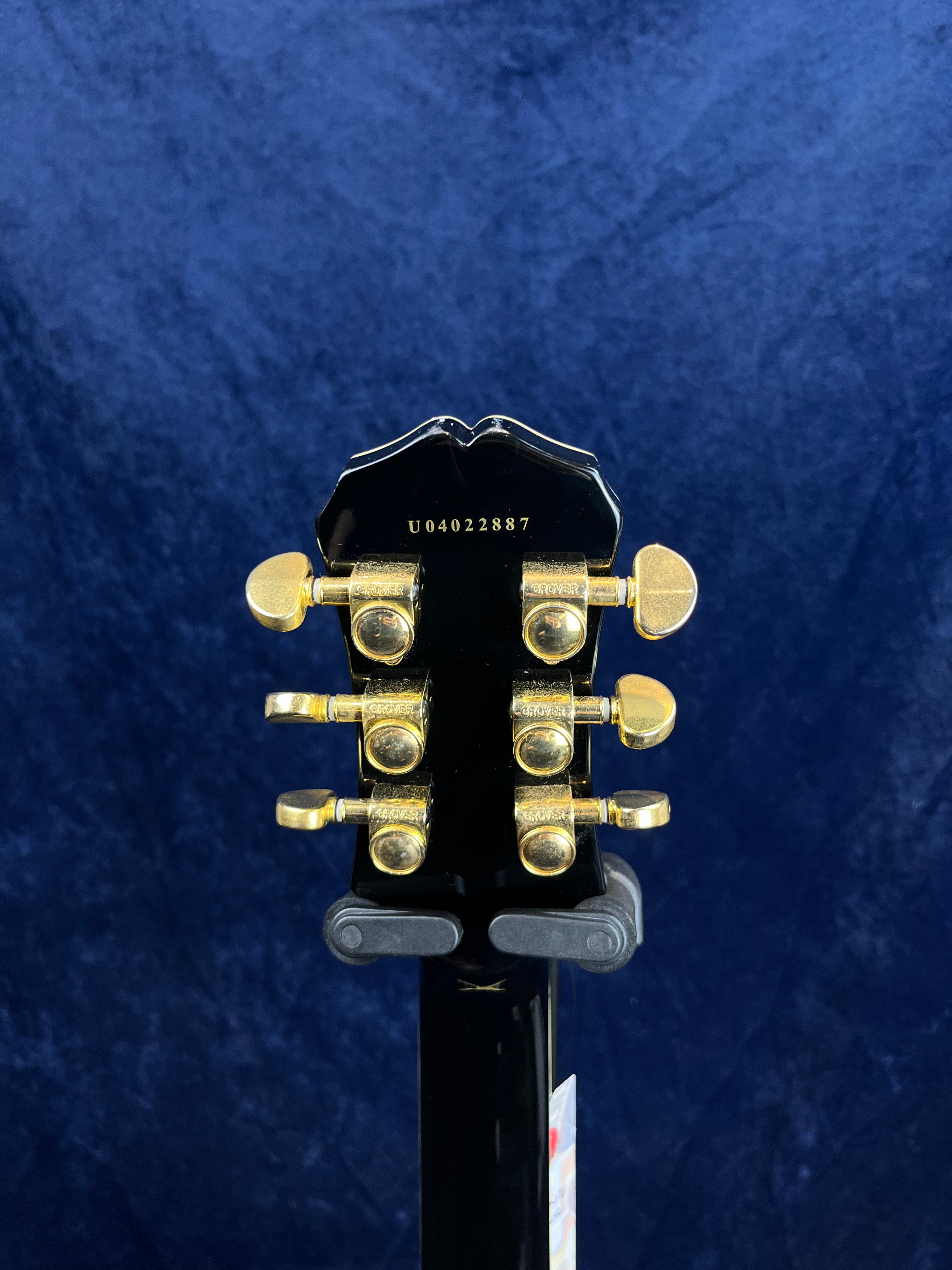 The back of a black electric guitar headstock with six gold tuning pegs and serial number U04022887, shown against a dark blue background.