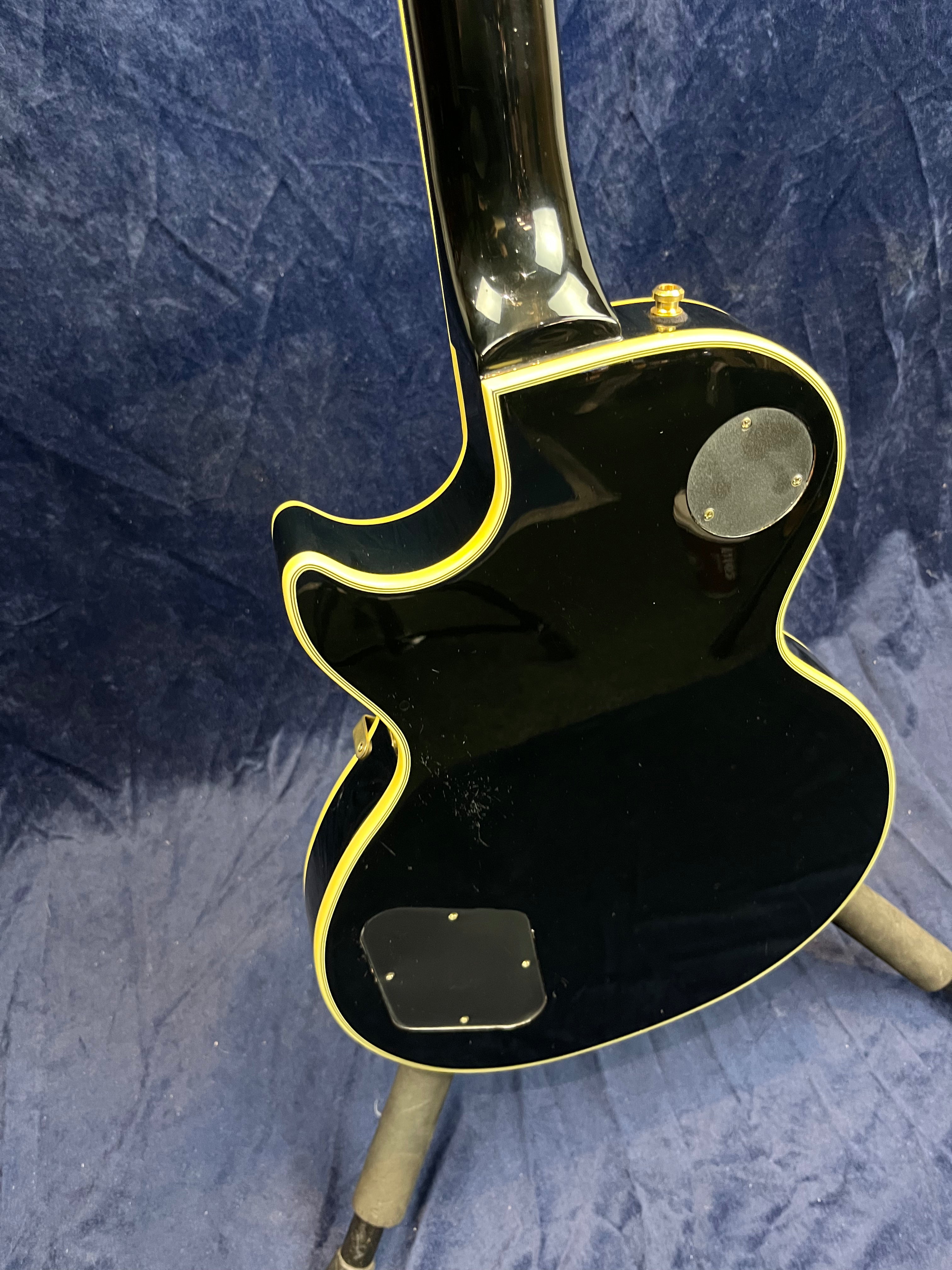 Glossy black electric guitar body with cream binding, two back cavity covers, and a single cutaway, shown from the rear on a stand against a dark backdrop.