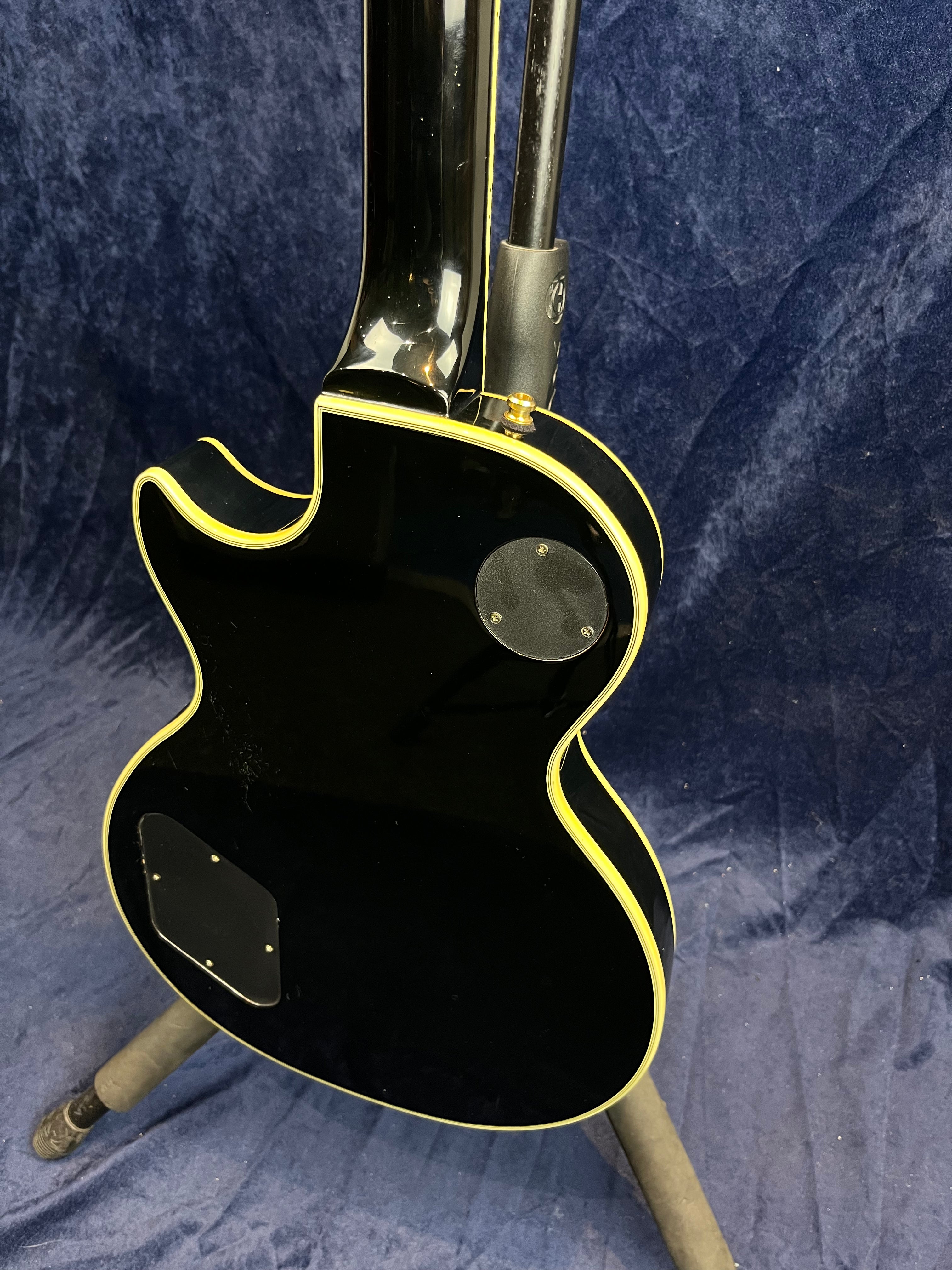 Black electric guitar with cream binding, viewed from the back on a stand, showing two rear access panels and a contoured single-cutaway body shape.