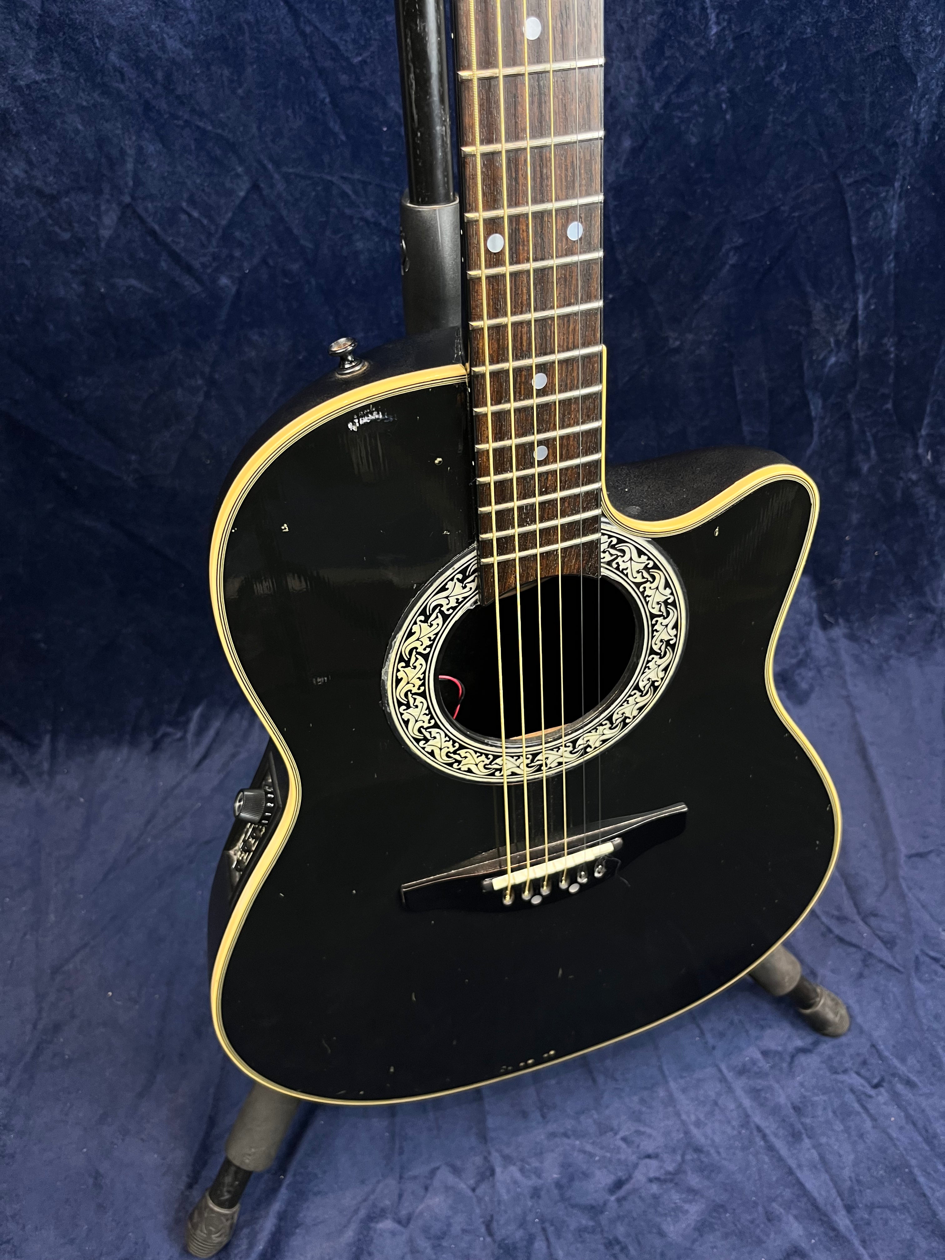 Black acoustic guitar with a decorative soundhole rosette, cream binding, and cutaway body design, shown upright on a stand against a dark background.