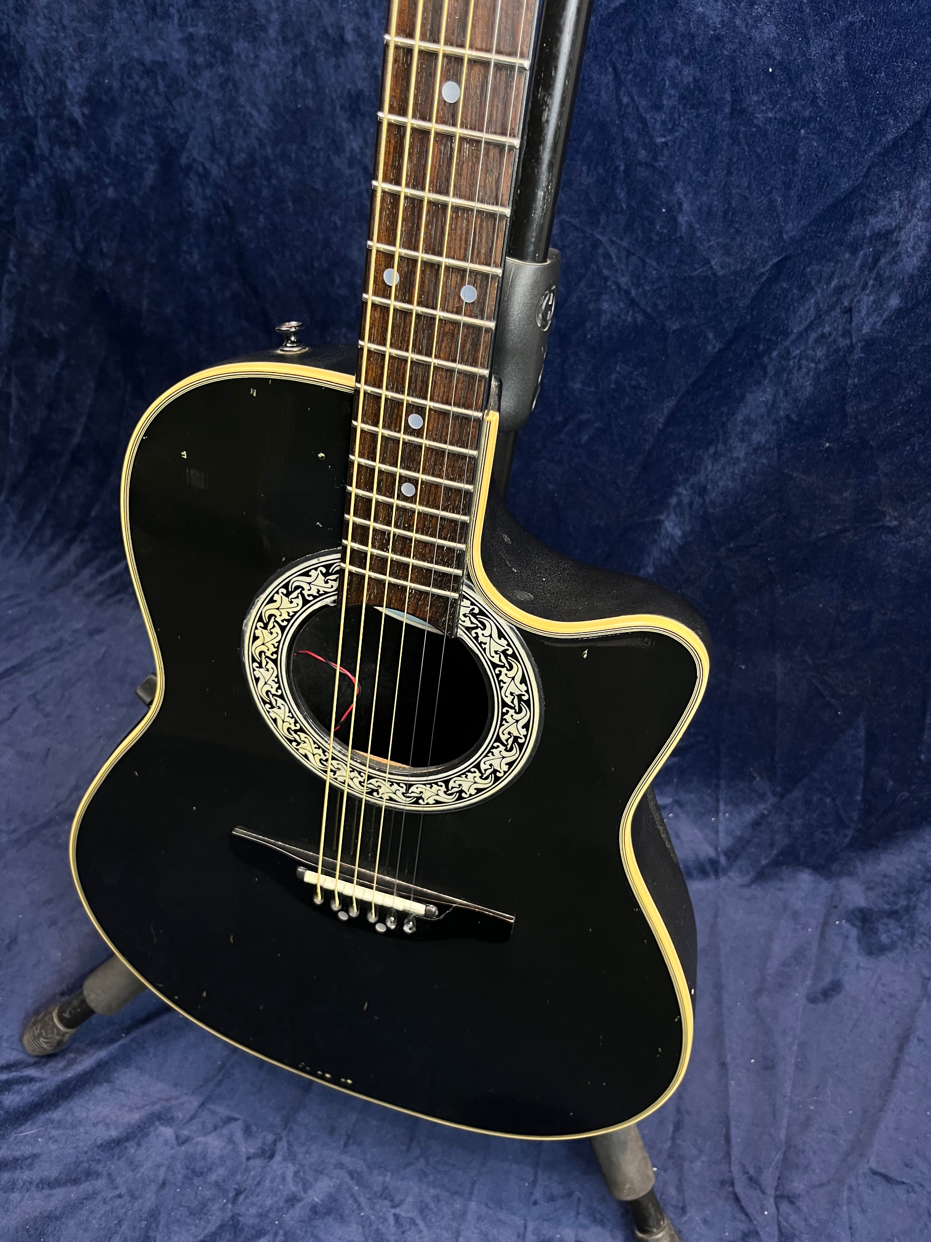Black acoustic guitar with a cutaway body, cream binding, ornate white rosette around the soundhole, and some visible scuffs on the lower front.