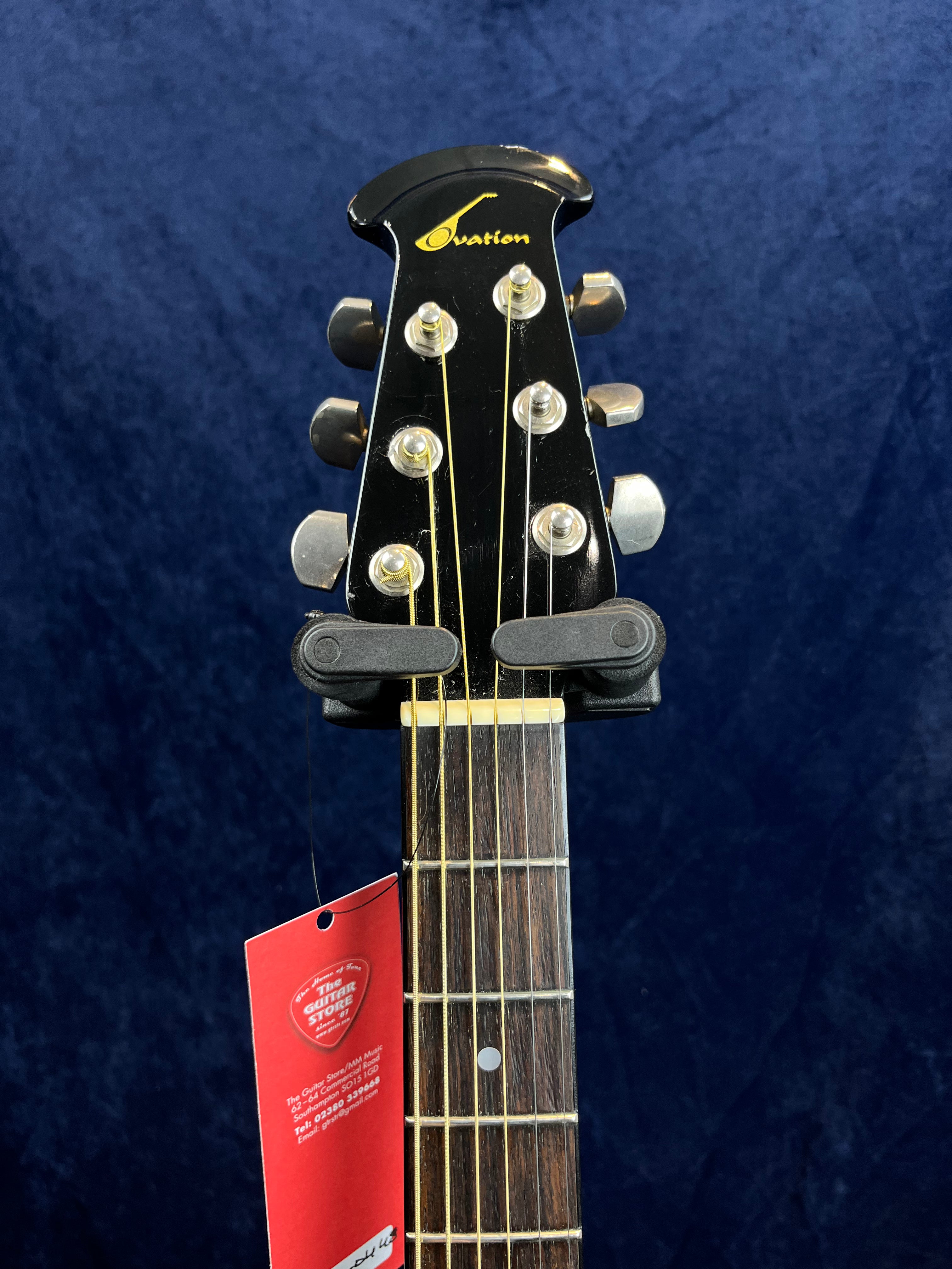 Black Ovation acoustic guitar headstock with six chrome tuners, yellow logo, rosewood fretboard, and a red tag hanging below the nut.