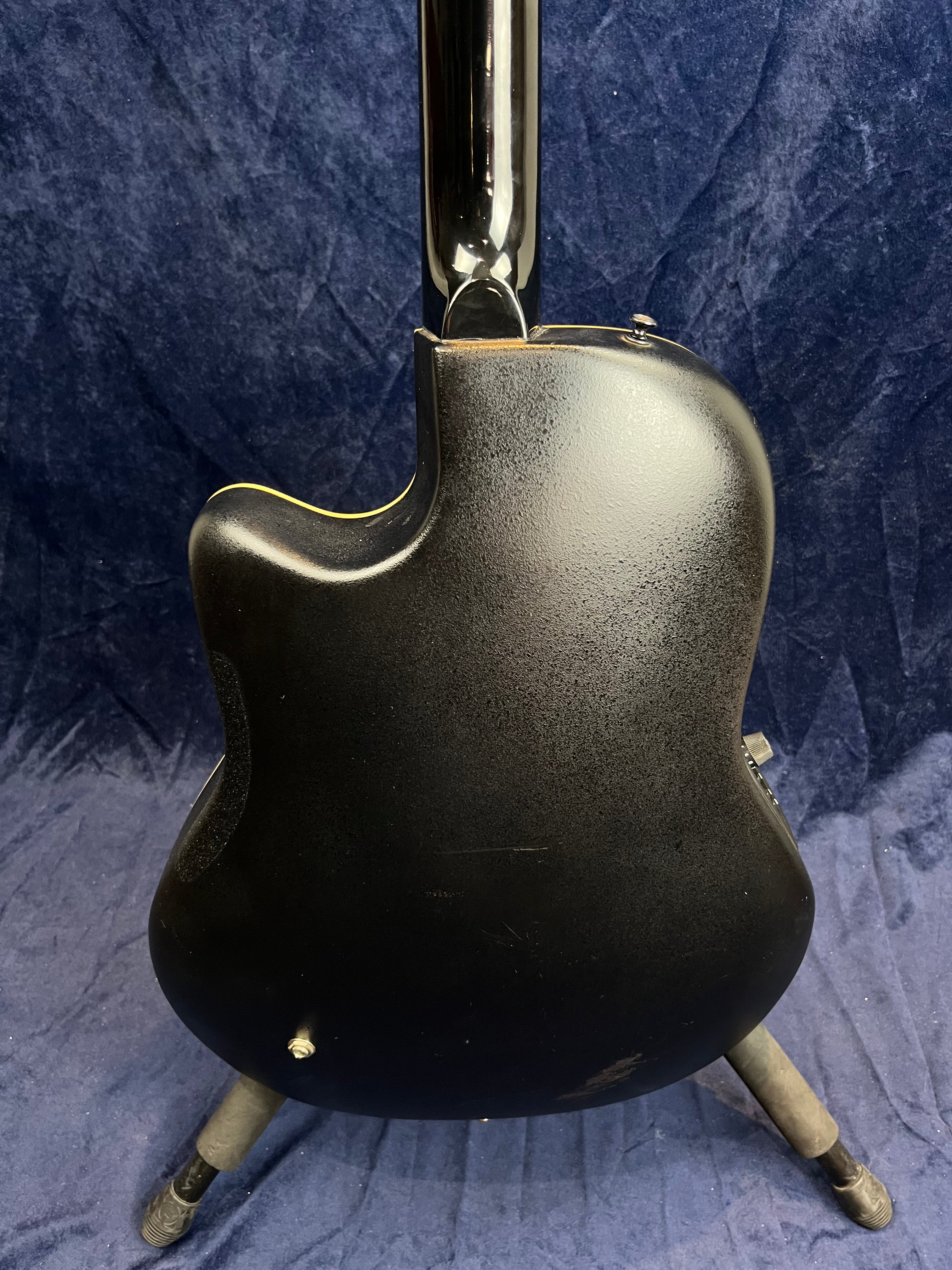 Black acoustic guitar shown from the back, with a cutaway design and visible minor scratches, resting on a stand against a dark blue background.