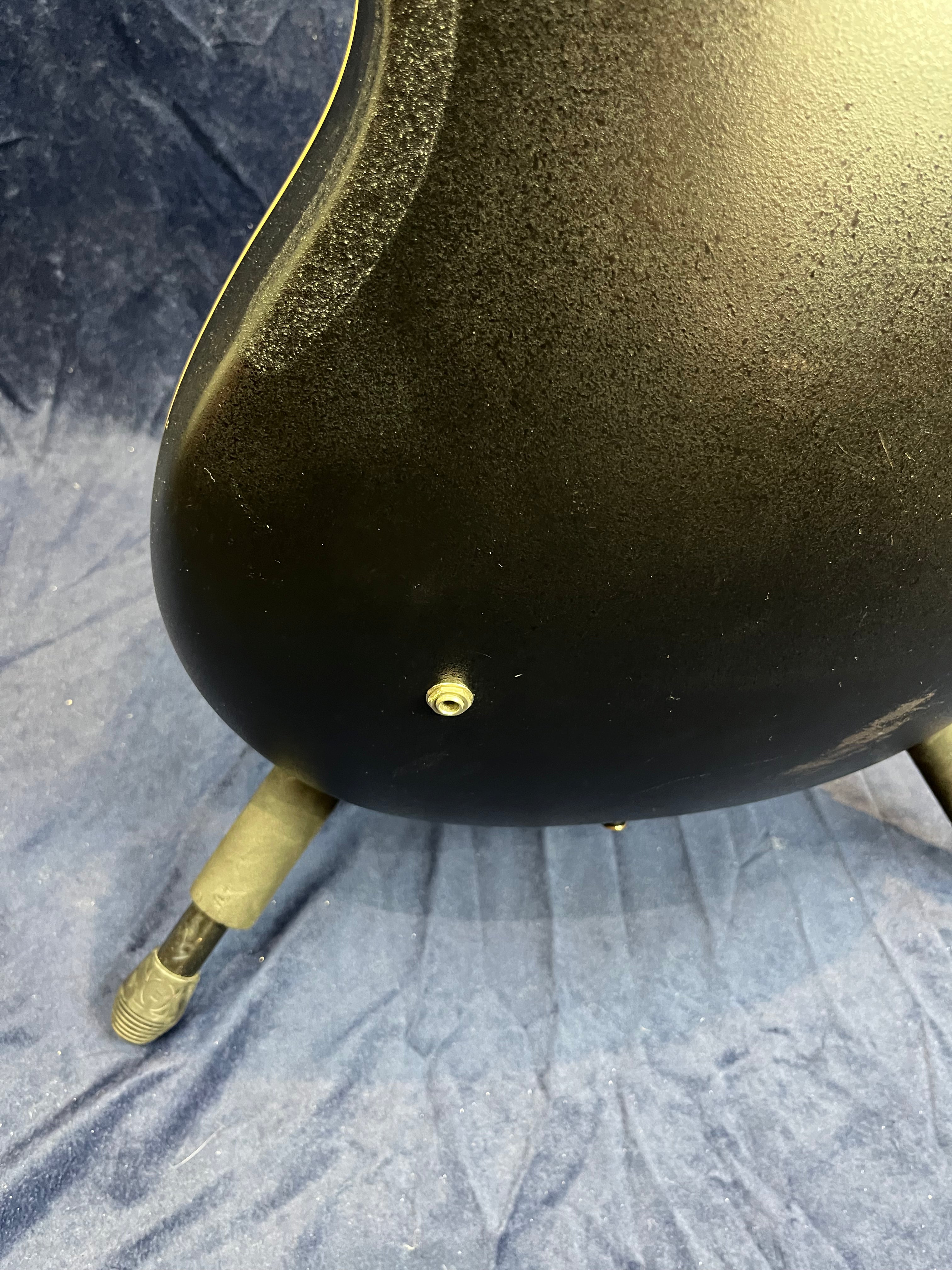 Black acoustic guitar body close-up showing the output jack on the side, resting on a padded stand over a blue cloth backdrop.