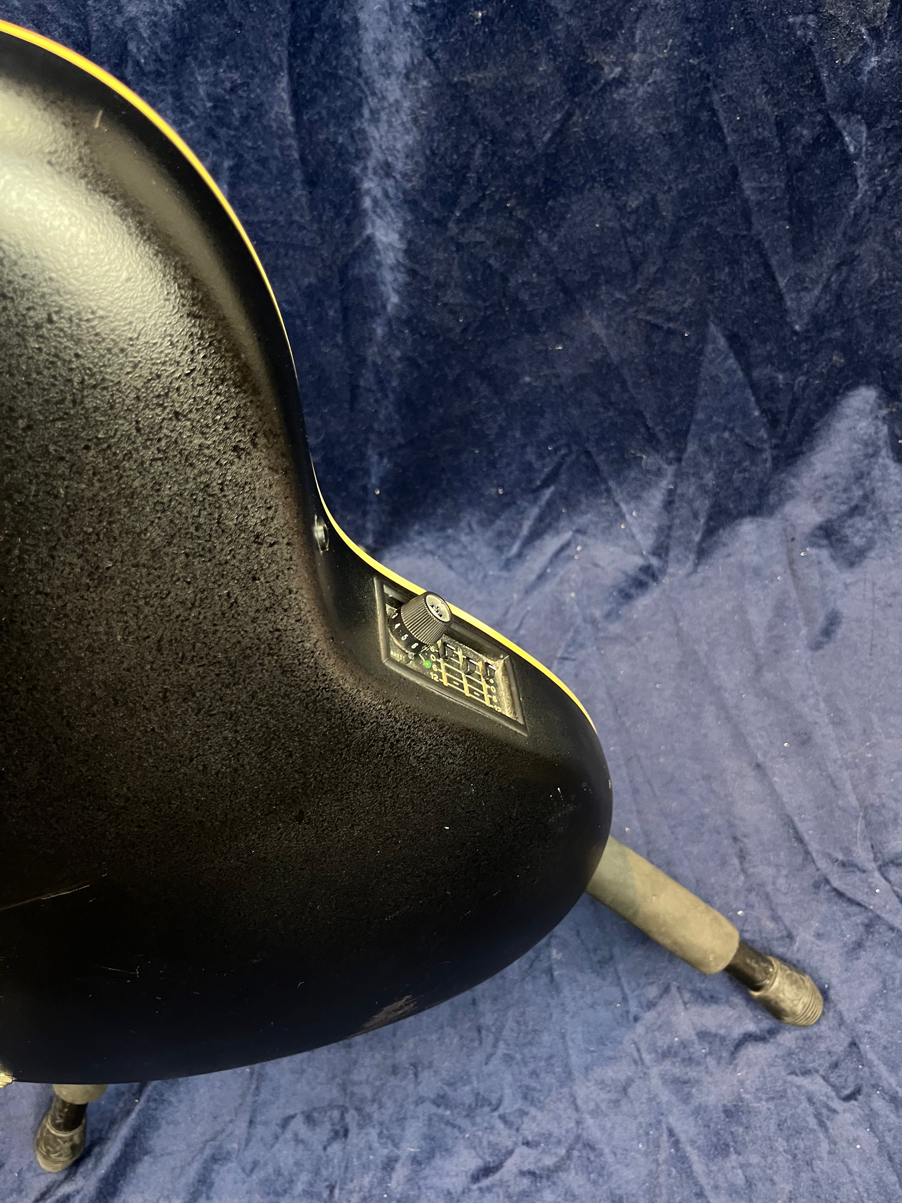 Black acoustic guitar back with a control panel embedded near the edge, resting on a stand against a textured blue fabric background.