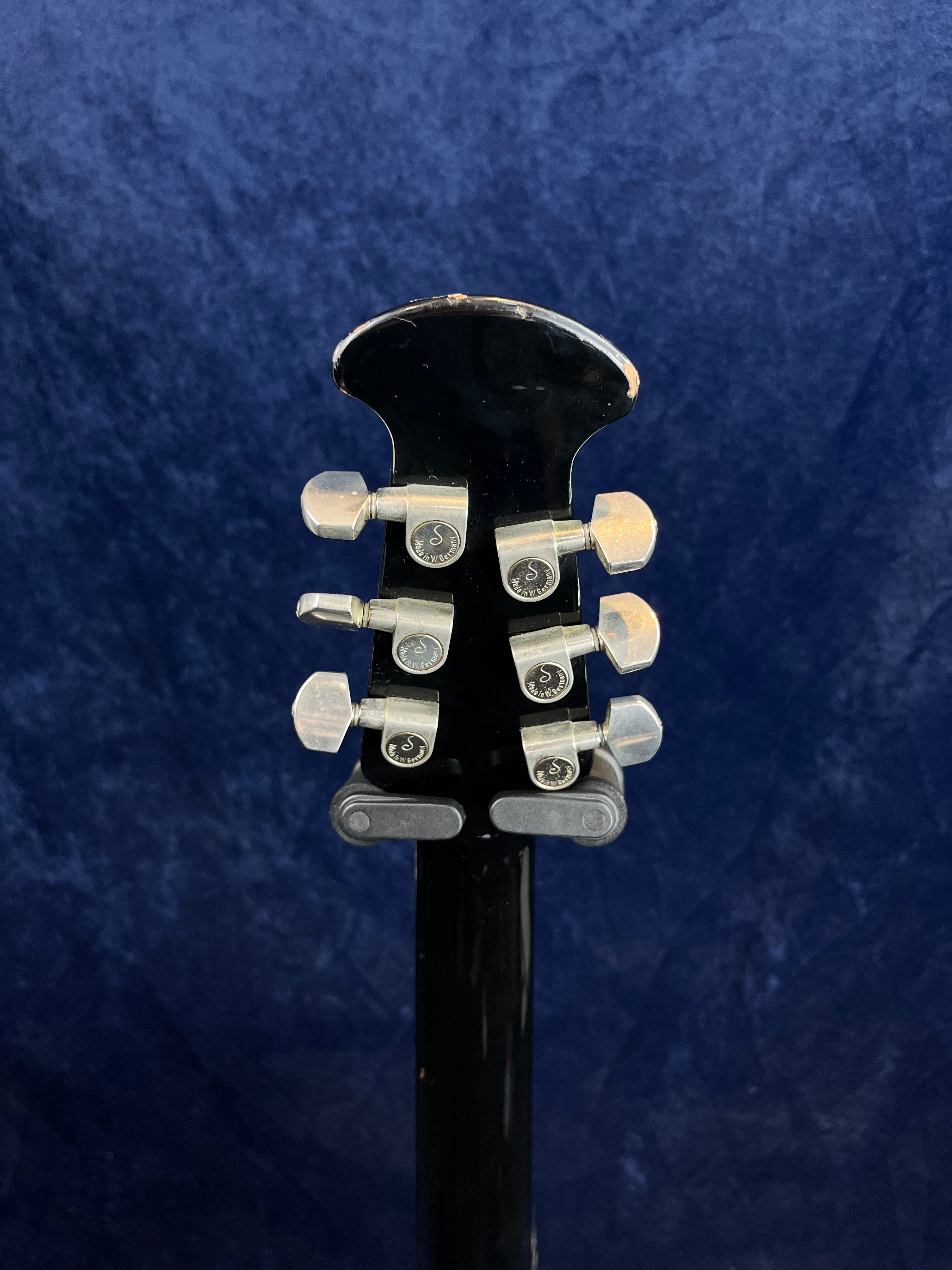 Back of a black acoustic guitar headstock with six silver tuning pegs, set against a dark blue textured background.