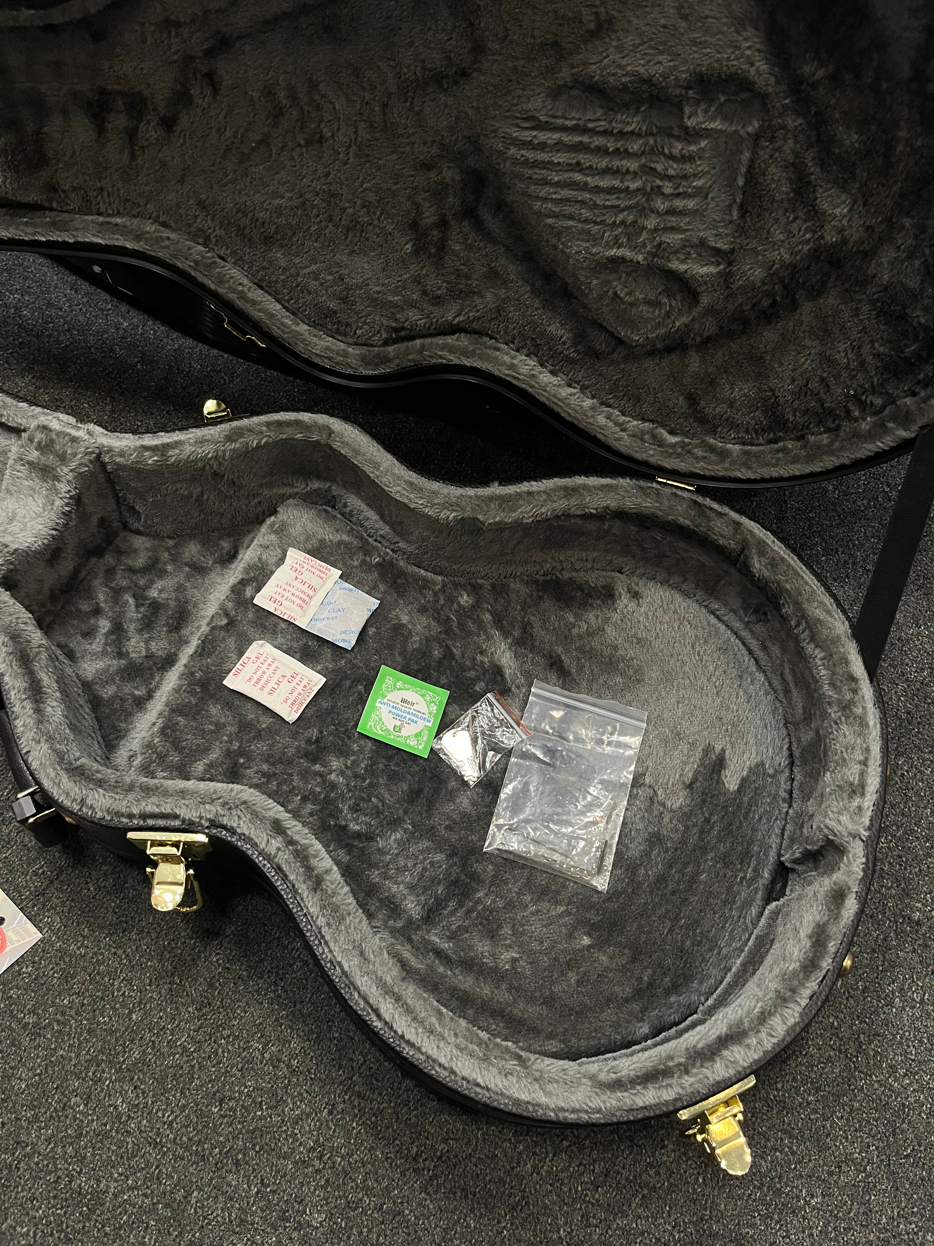 Open electric guitar case with gray plush lining, showing spare strings, silica gel packets, and small clear bags with hardware inside.