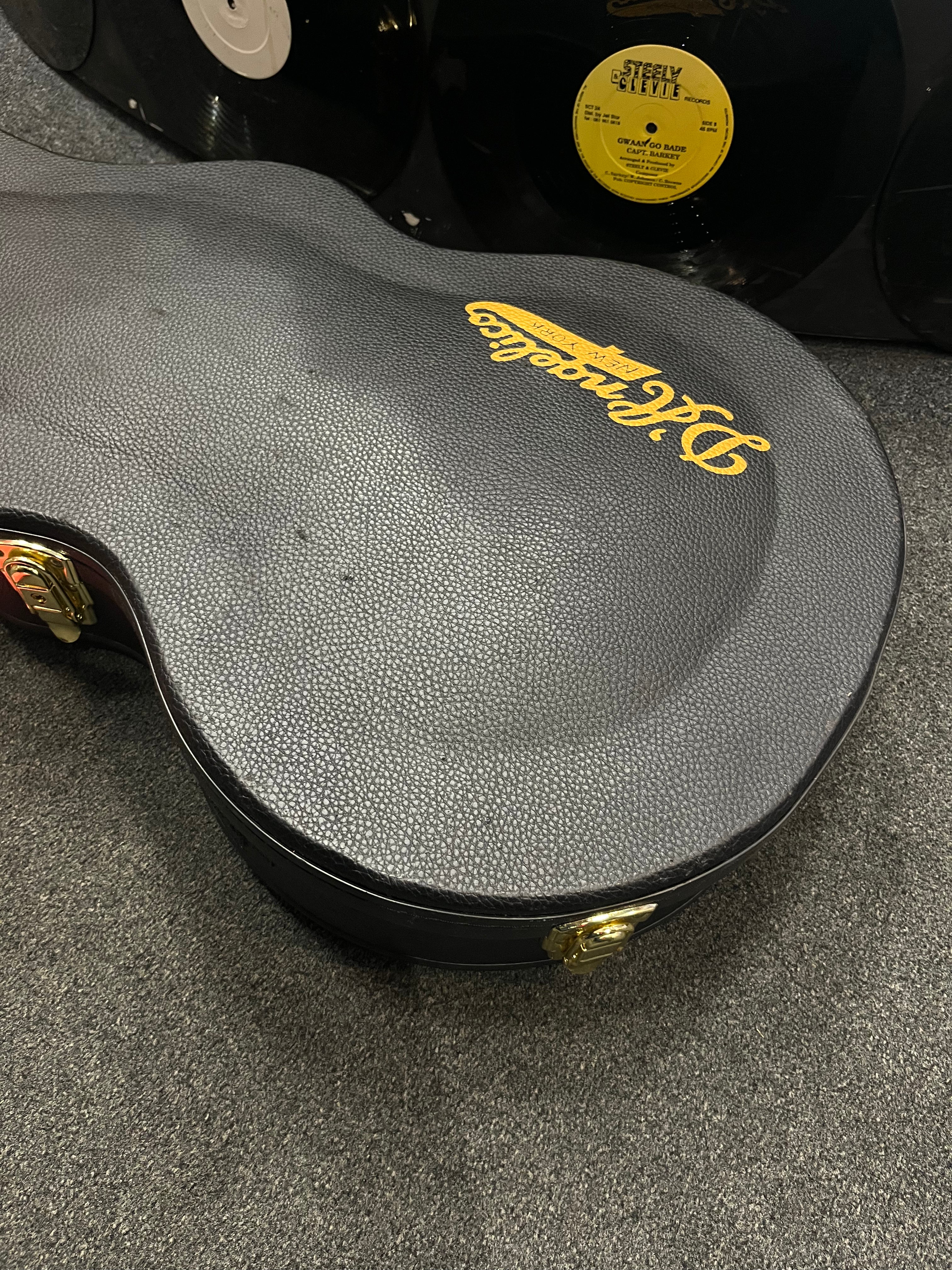 A black textured electric guitar case with gold hardware and a yellow Epiphone logo sits on carpet, near vinyl records leaning against a wall.