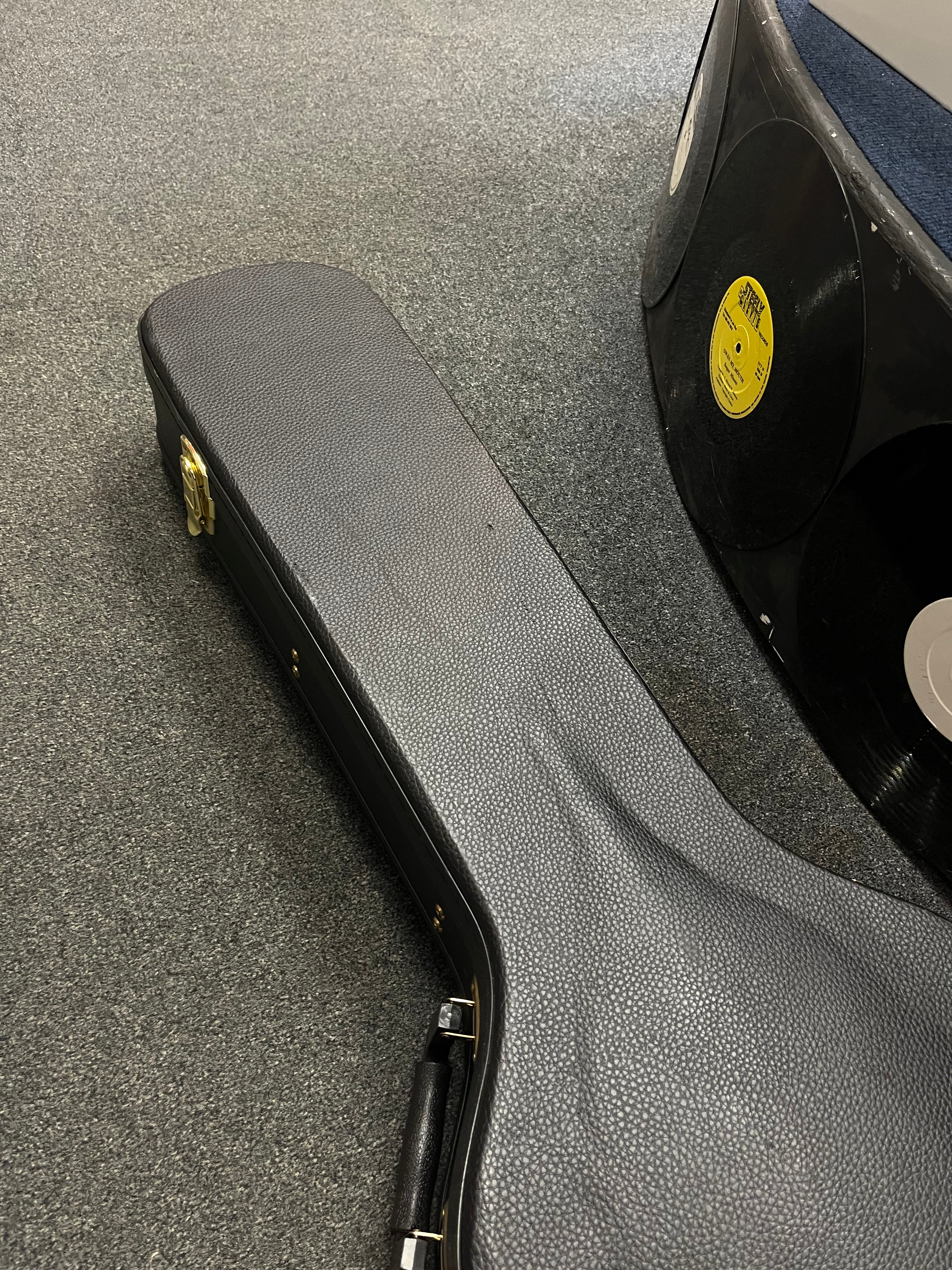 A black textured electric guitar case with a handle rests on carpet, next to an object decorated with vinyl records.