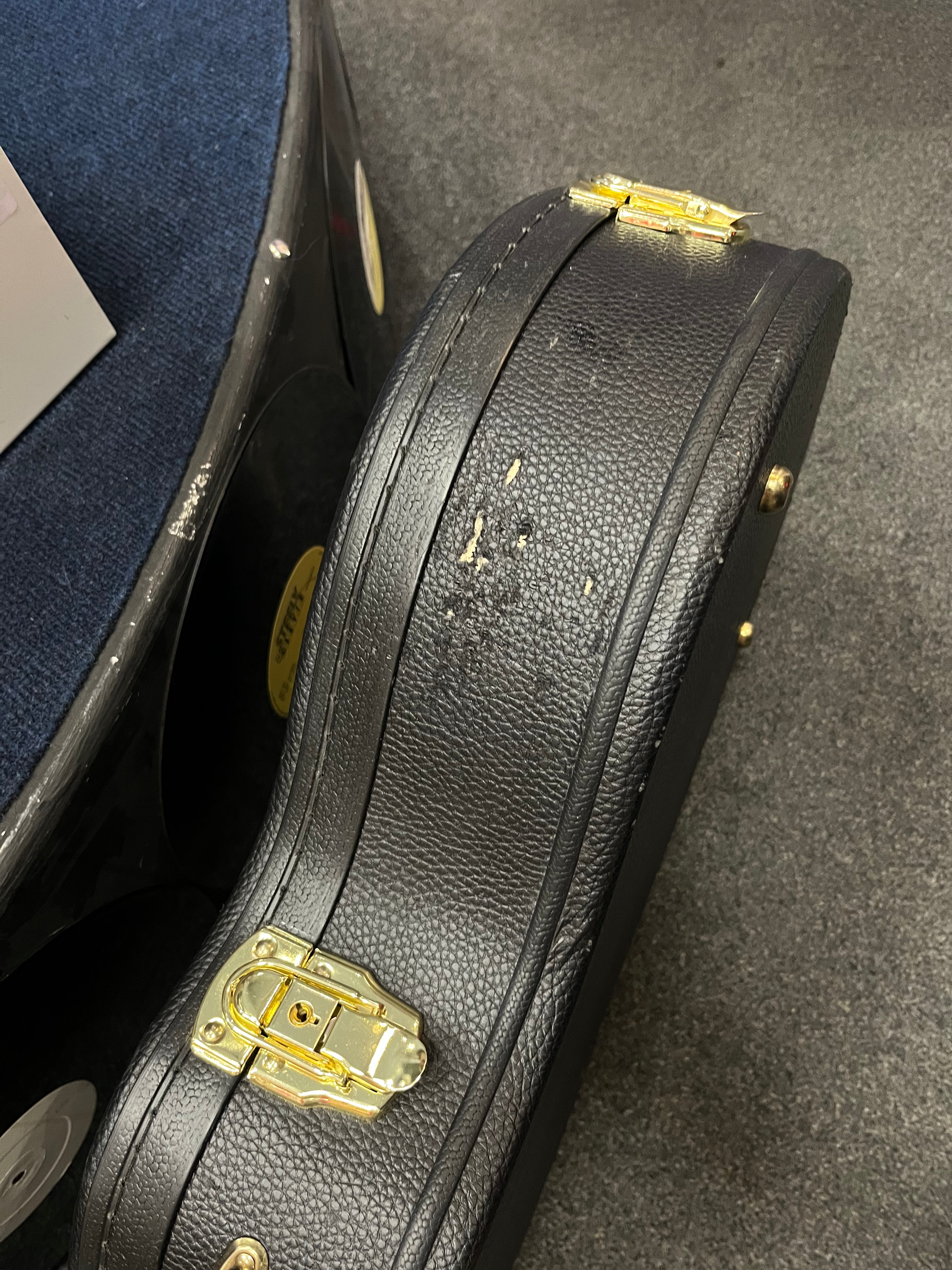 Black textured electric guitar case with gold latches and some visible scuffs and wear on the surface, resting on a dark carpeted floor.