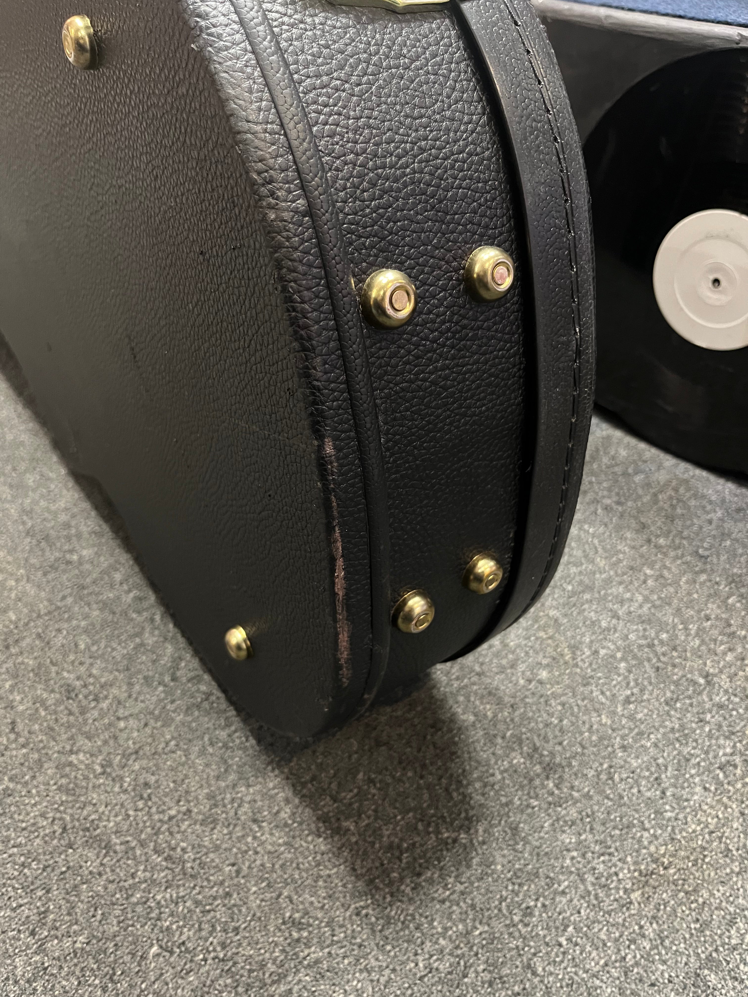 Black textured hard guitar case with brass feet, slight scuff on the edge, part of a vinyl record visible in the background.