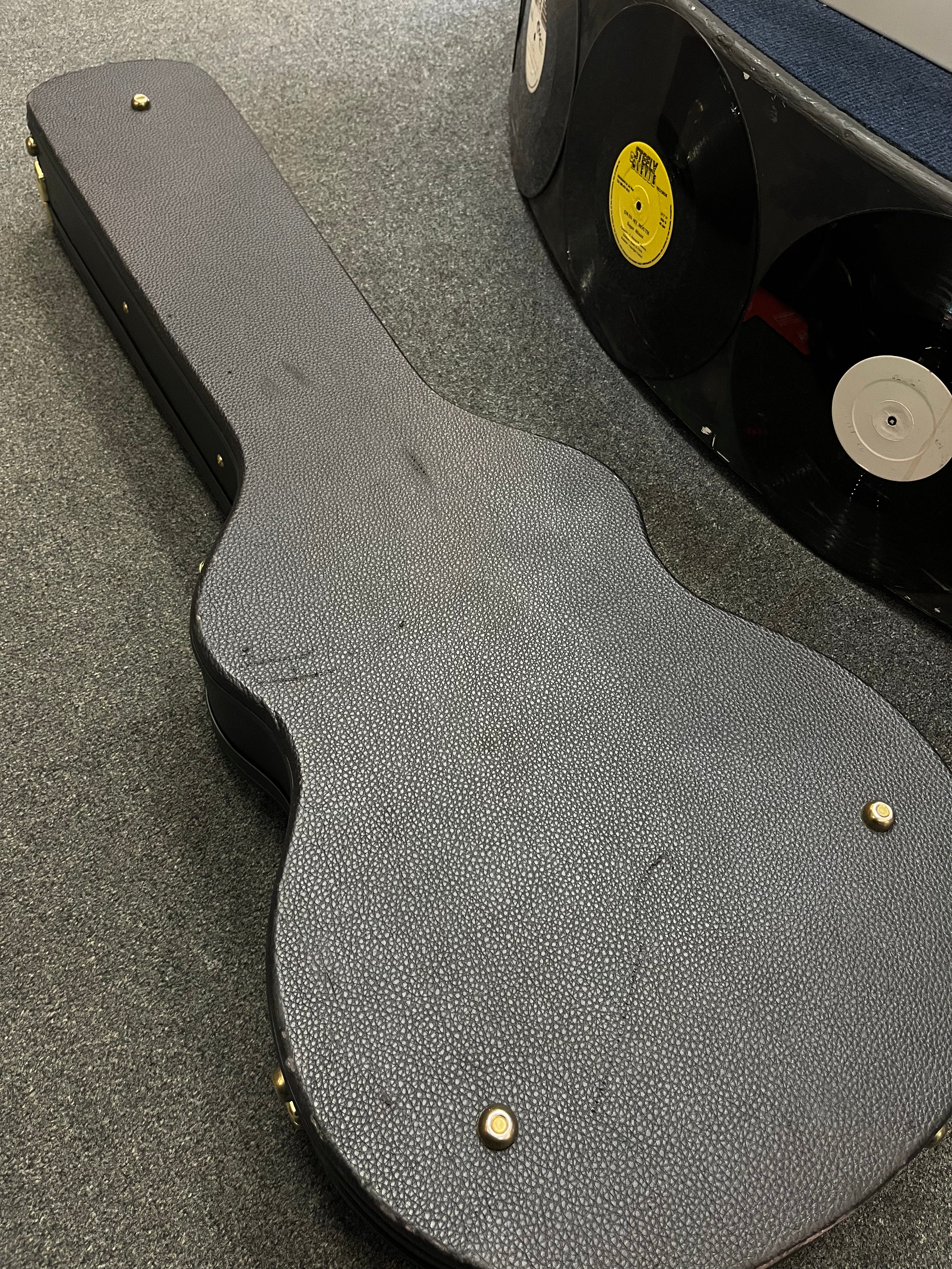 A closed, textured black electric guitar case with metal studs lies on carpet next to furniture decorated with vinyl records.