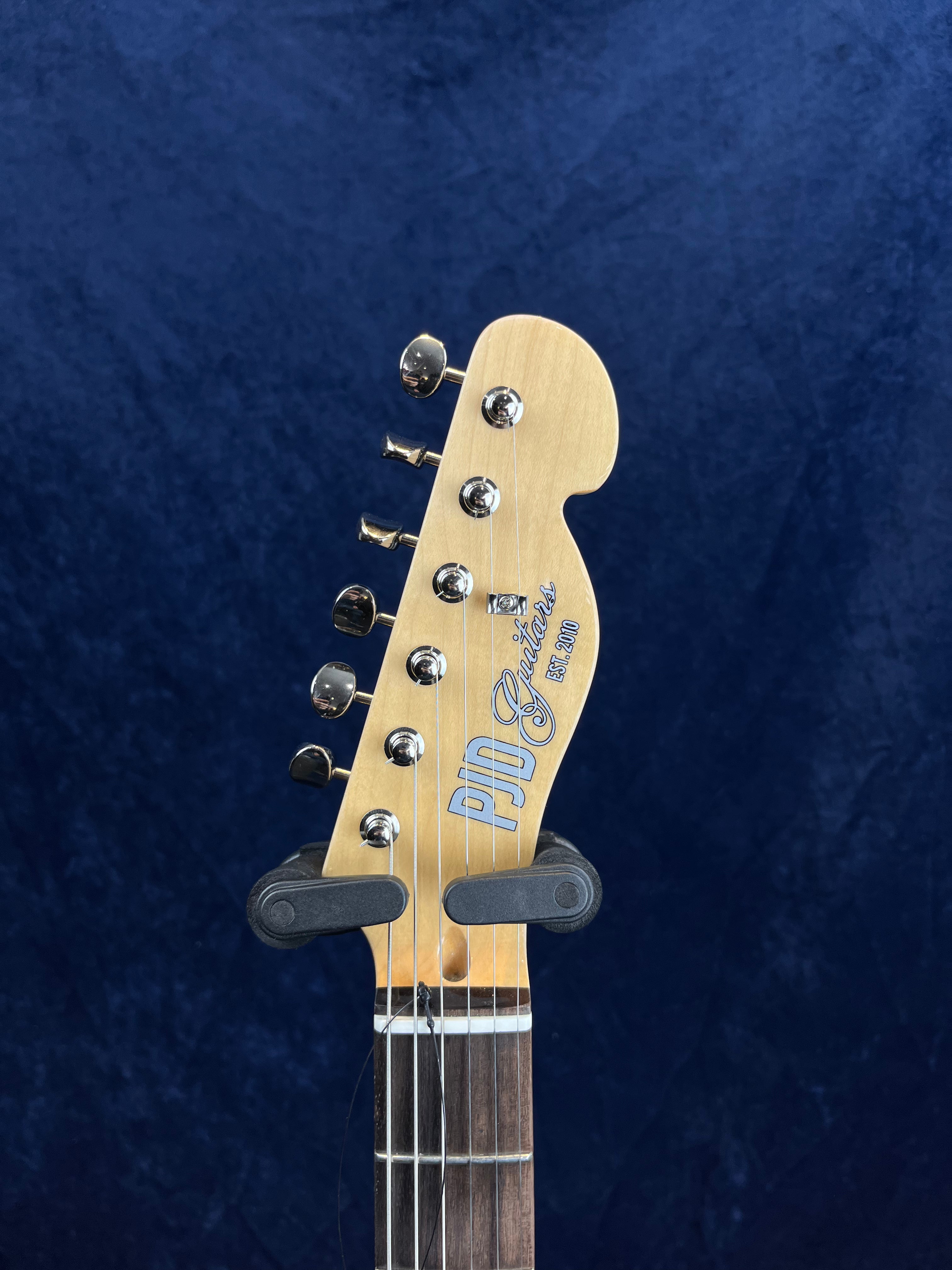 Light wood electric guitar headstock with six silver tuning pegs, "PJD Guitars EST. 2010" logo, and a dark fretboard against a blue background.