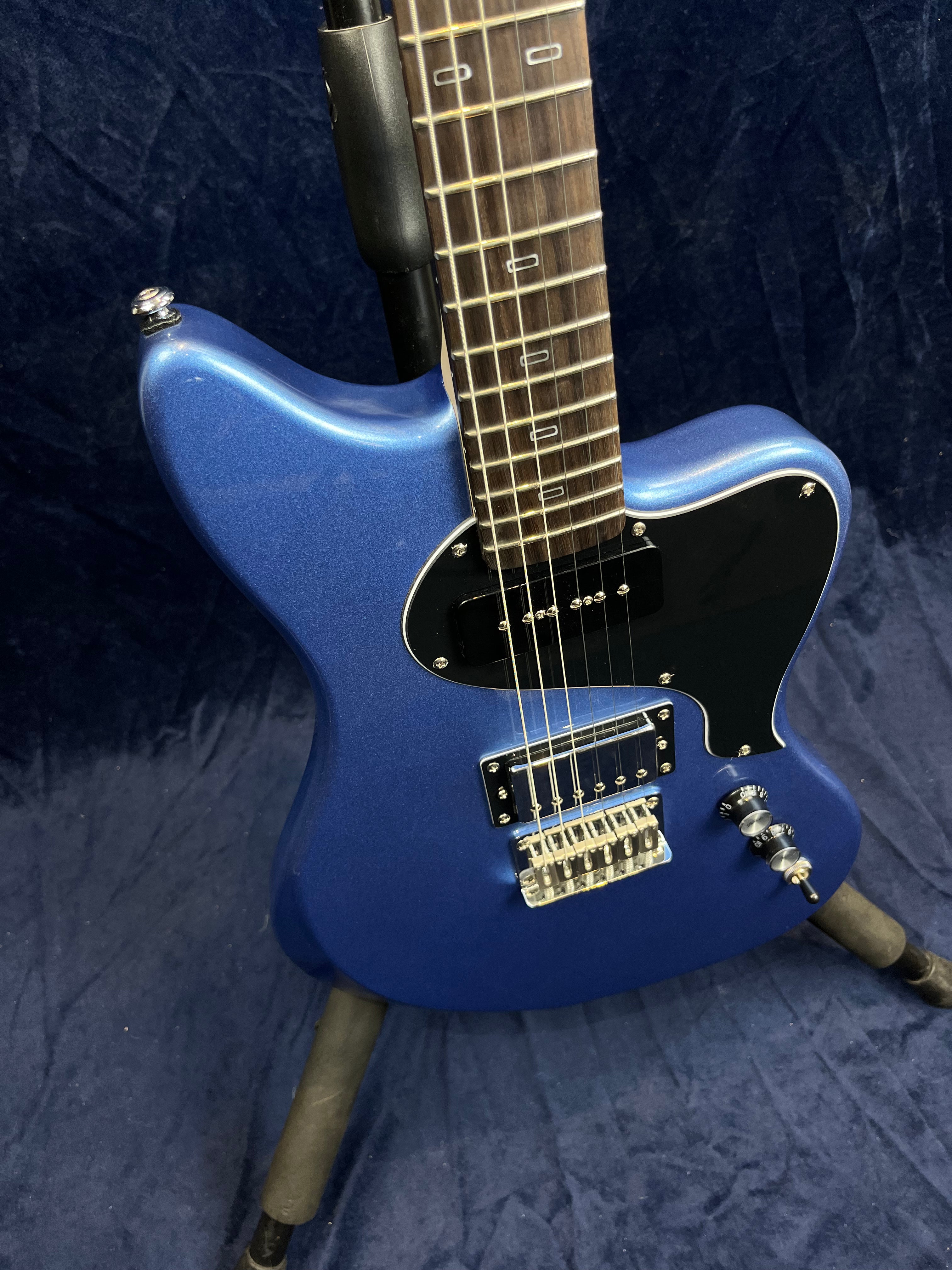 Metallic blue electric guitar with black pickguard, two pickups, three control knobs, and a switch, shown on a stand against a dark background.