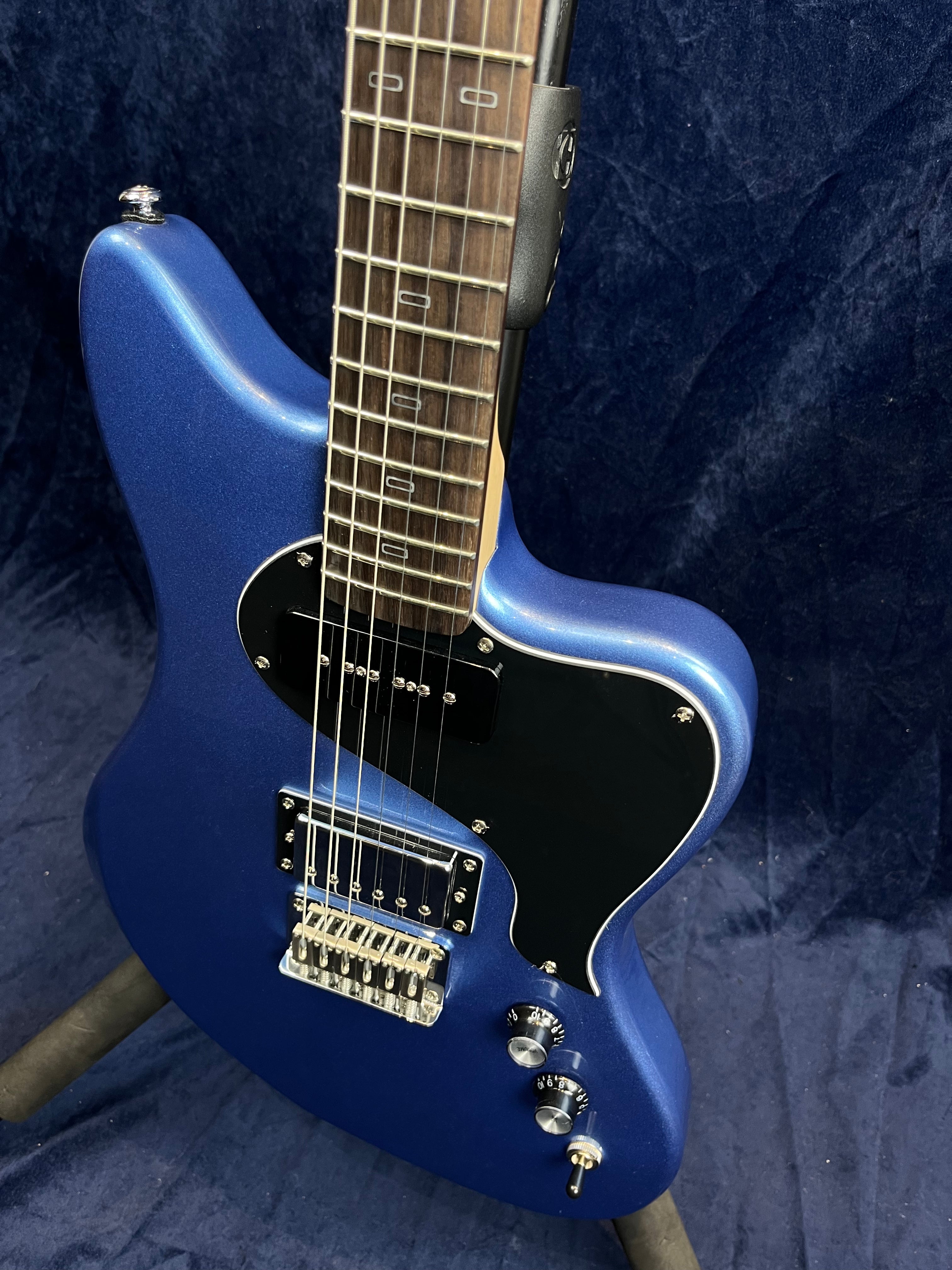 Metallic blue electric guitar with a black pickguard, two control knobs, a toggle switch, and a chrome bridge, shown on a stand.