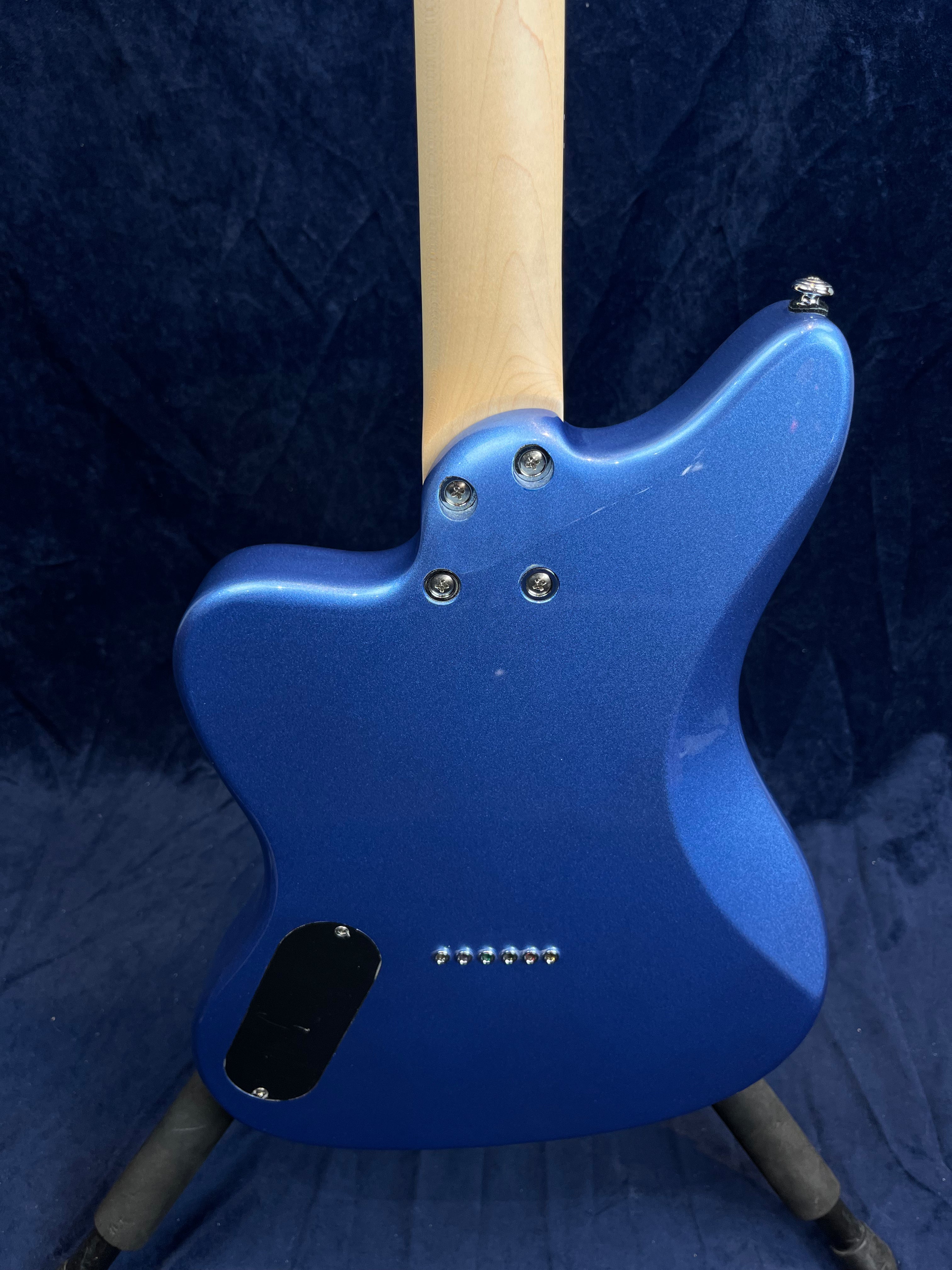 Metallic blue electric guitar viewed from the back, showing a light wood neck, four bolt neck plate, string ferrules, and an access panel on the body.