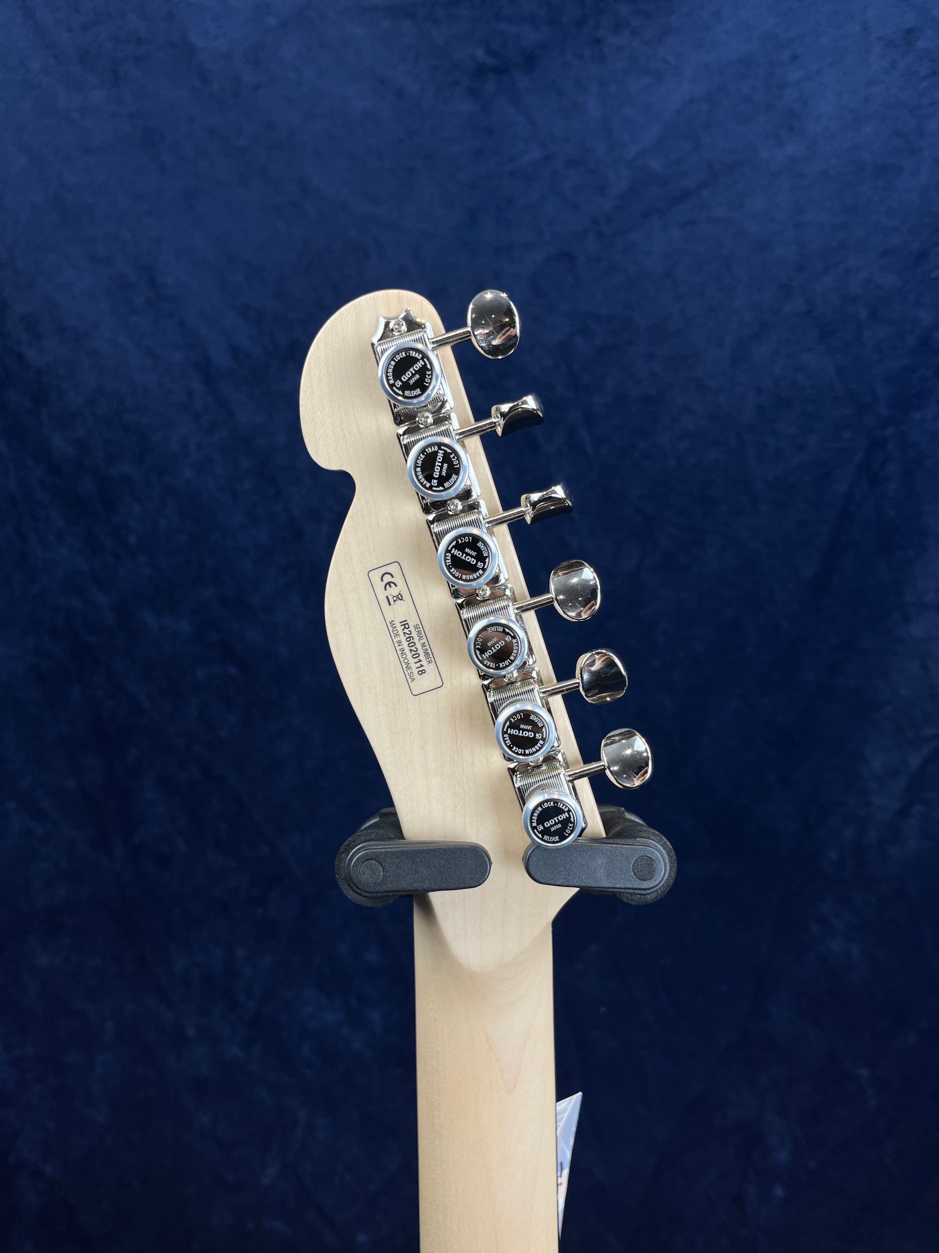 Six chrome tuning pegs line the back of a light wood electric guitar headstock against a dark blue background; the guitar is held by a wall hanger.