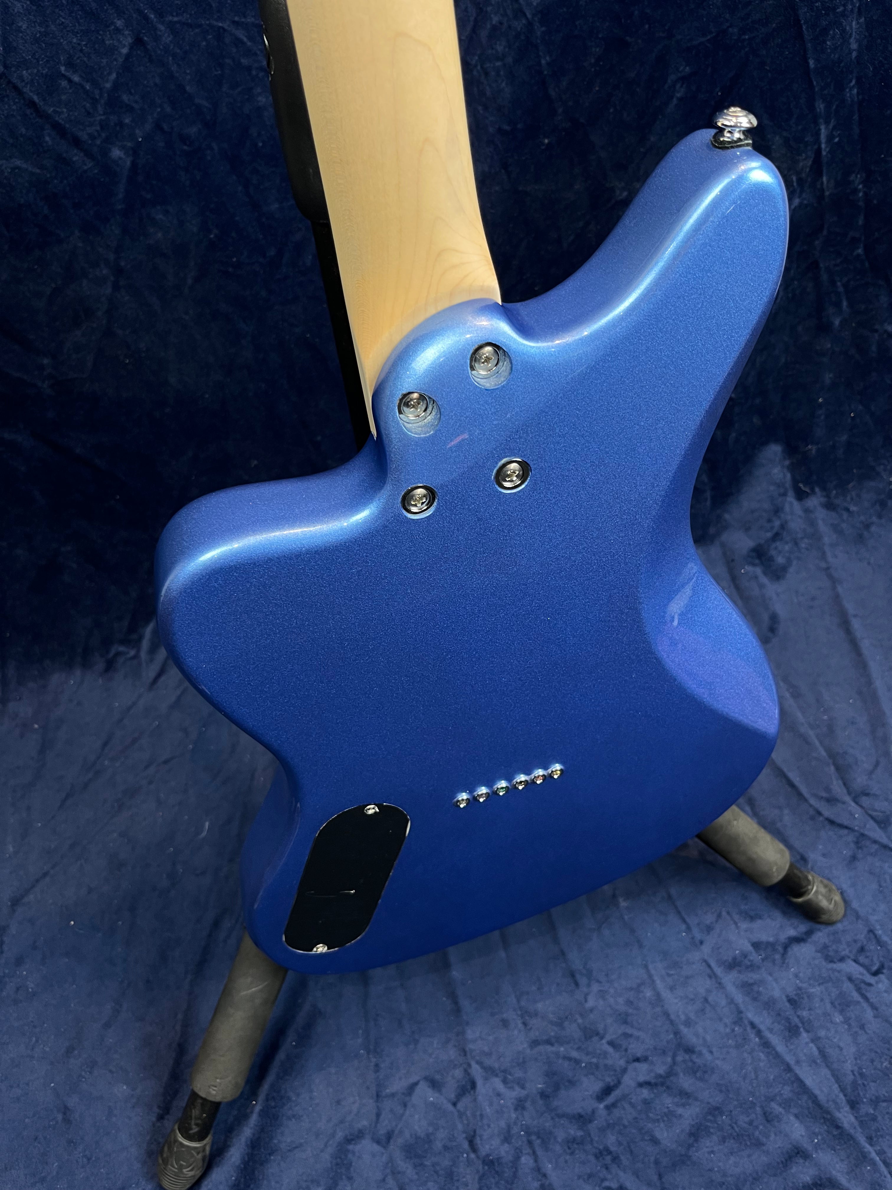 Blue electric guitar seen from the back, showing a bolt-on natural wood neck, string ferrules, and a small black electronics cover on a stand.